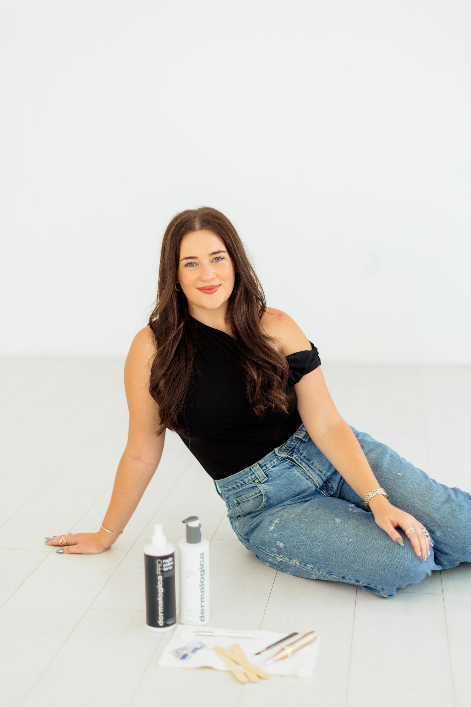 Woman in black top and jeans sits on white floor with hair coloring products and brushes beside her.