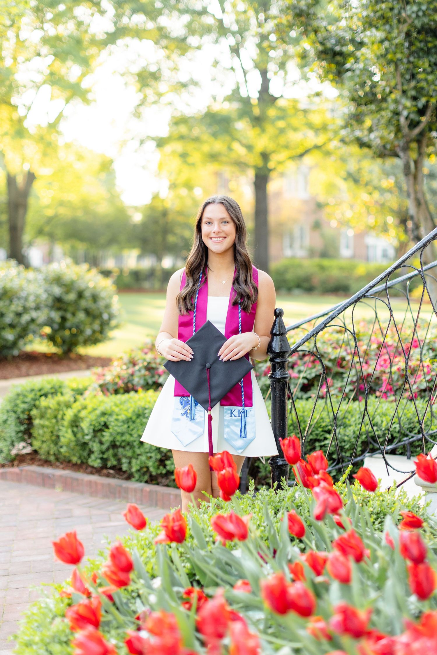 Graduate in white dress with diploma standing by red tulip garden.