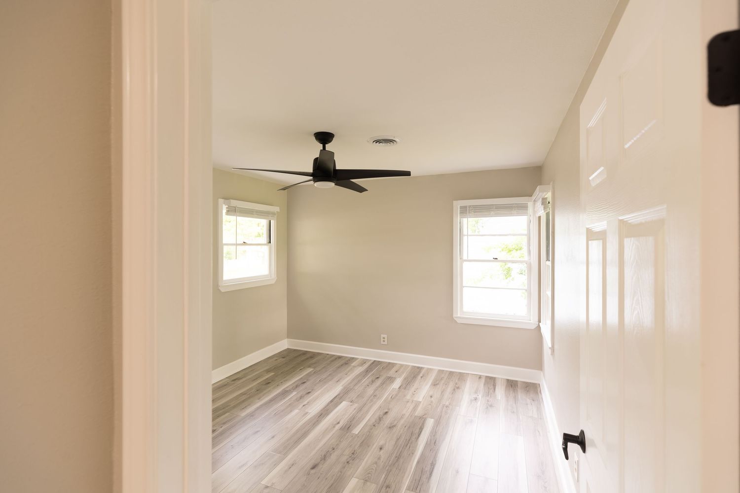 Empty bedroom with light wood floors, ceiling fan, two windows, and neutral gray walls in a modern home.