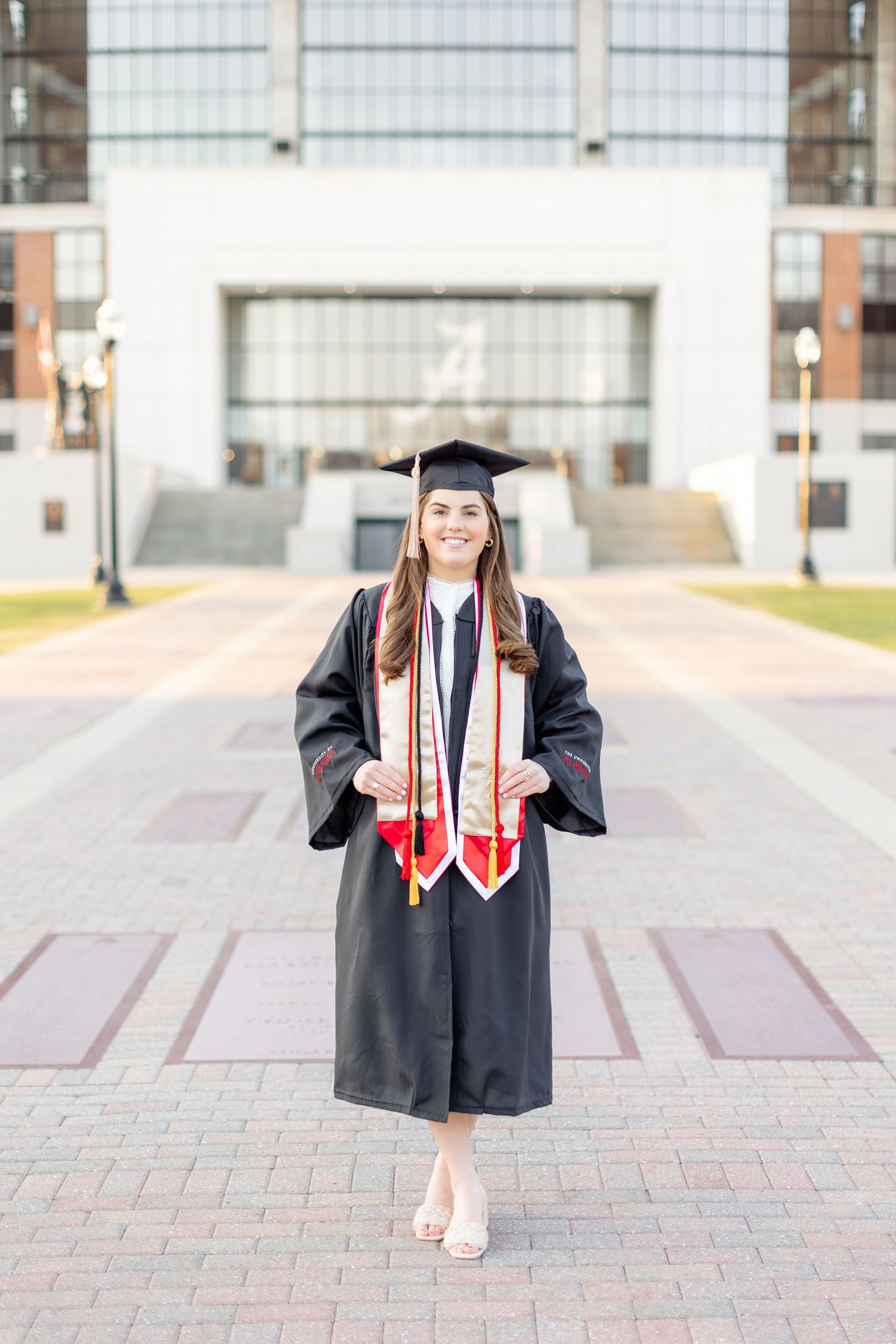 Graduate in black cap and gown with red and white stoles poses on brick pathway in front of campus building.