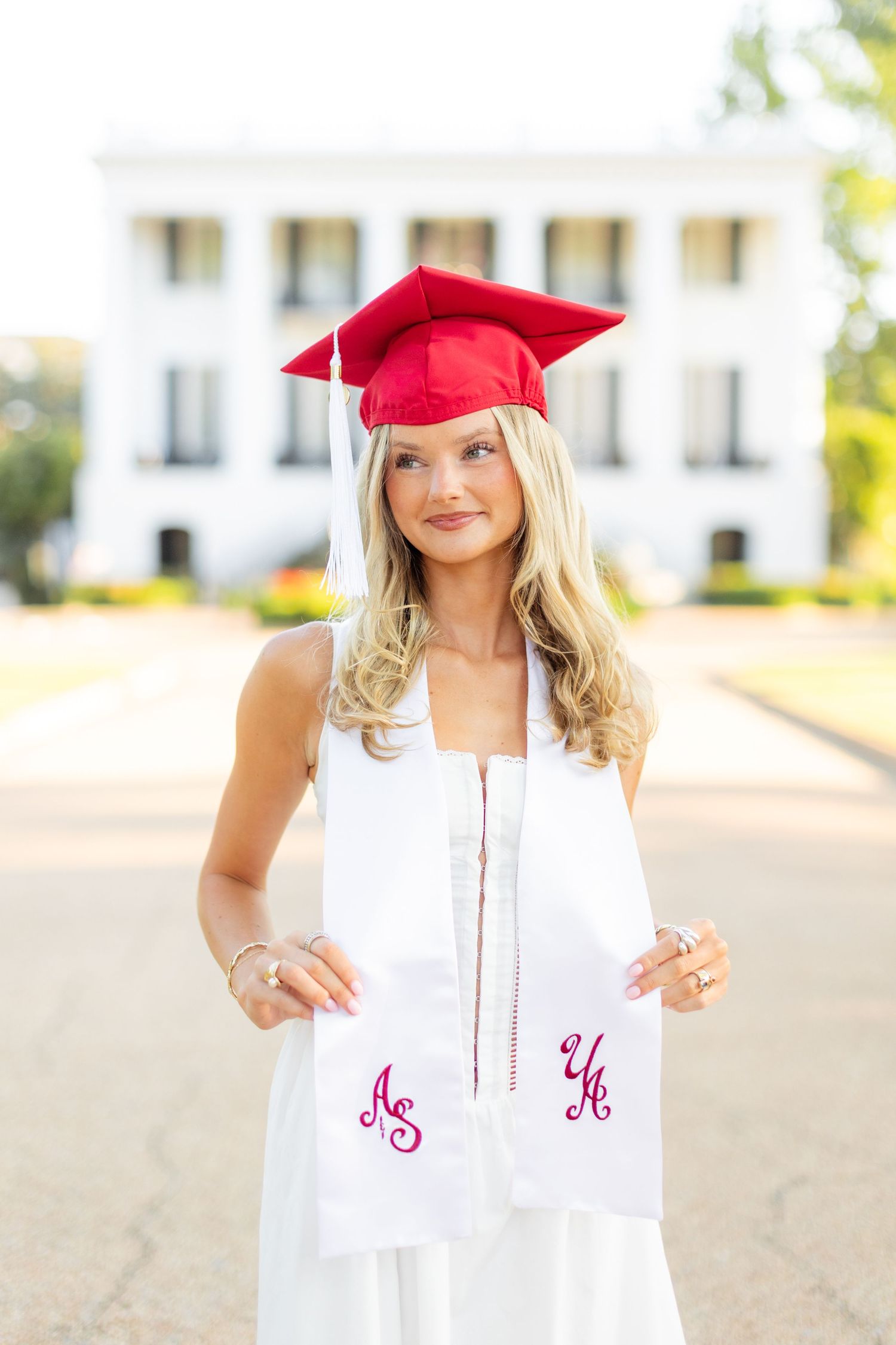 Graduate in white dress and red cap posing in front of university building.