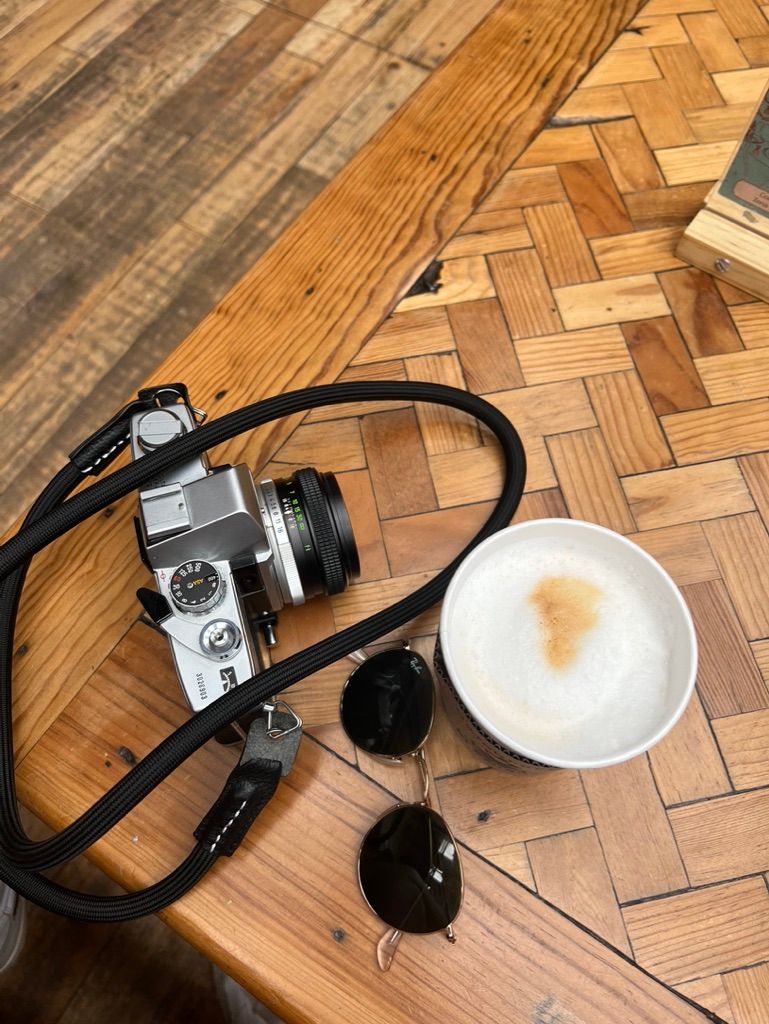 A vintage camera and sunglasses arranged next to a latte on a herringbone wooden floor.