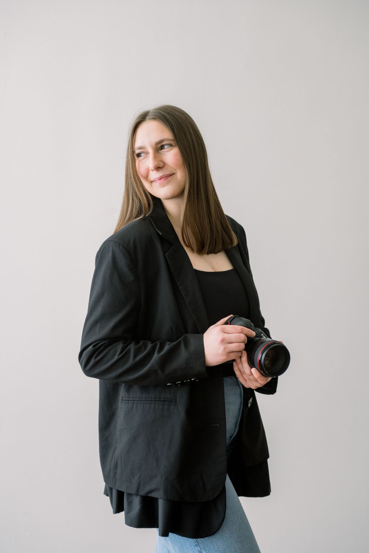 Professional photographer in a black blazer and jeans holds a camera while posing against a light gray background.