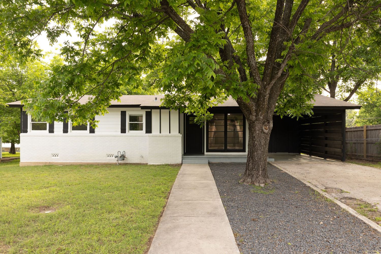White ranch-style home with large shade trees, gravel driveway, and covered carport in a suburban neighborhood.