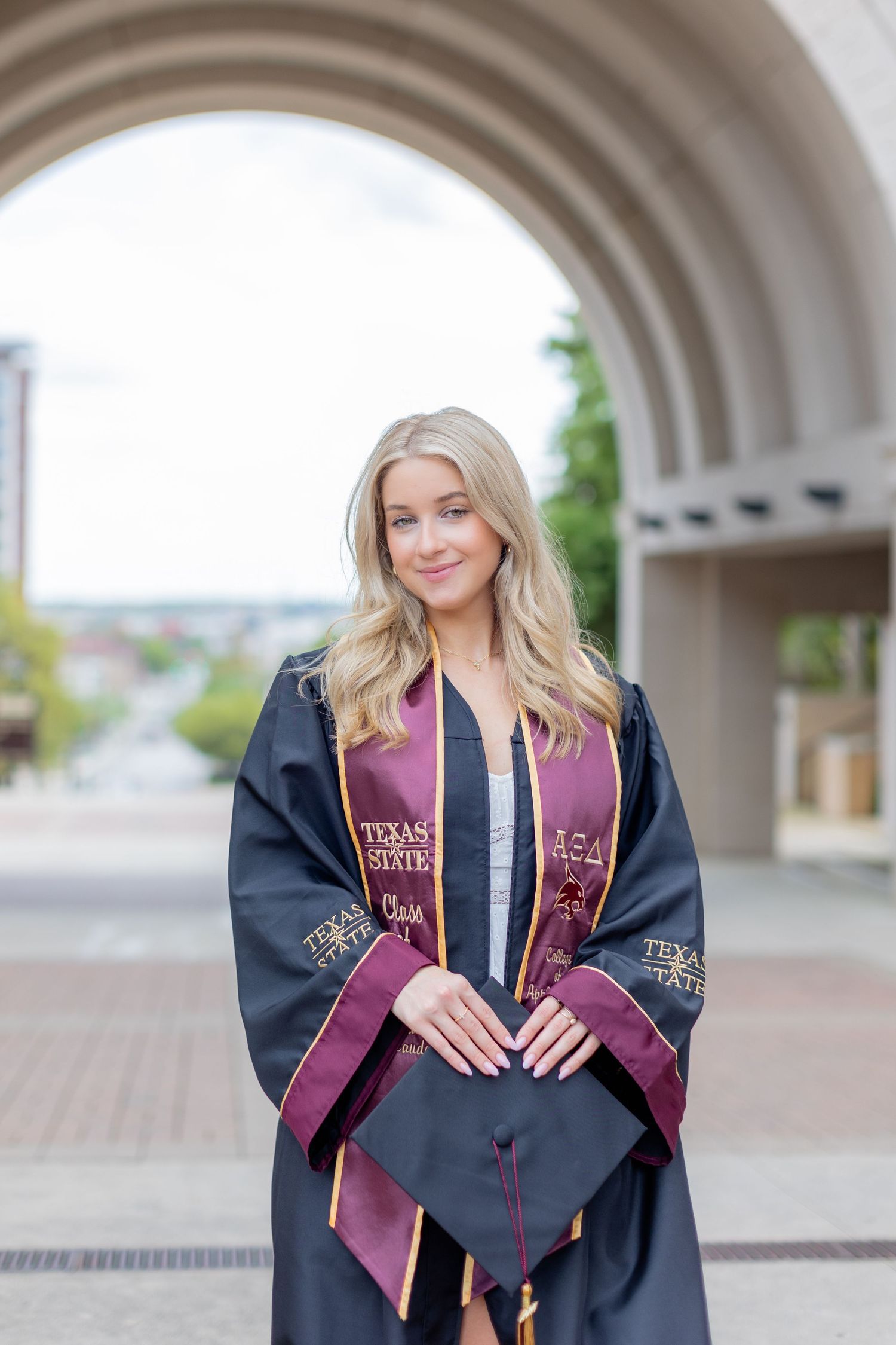 A graduation portrait showing traditional cap and gown with maroon accents under an architectural arch.