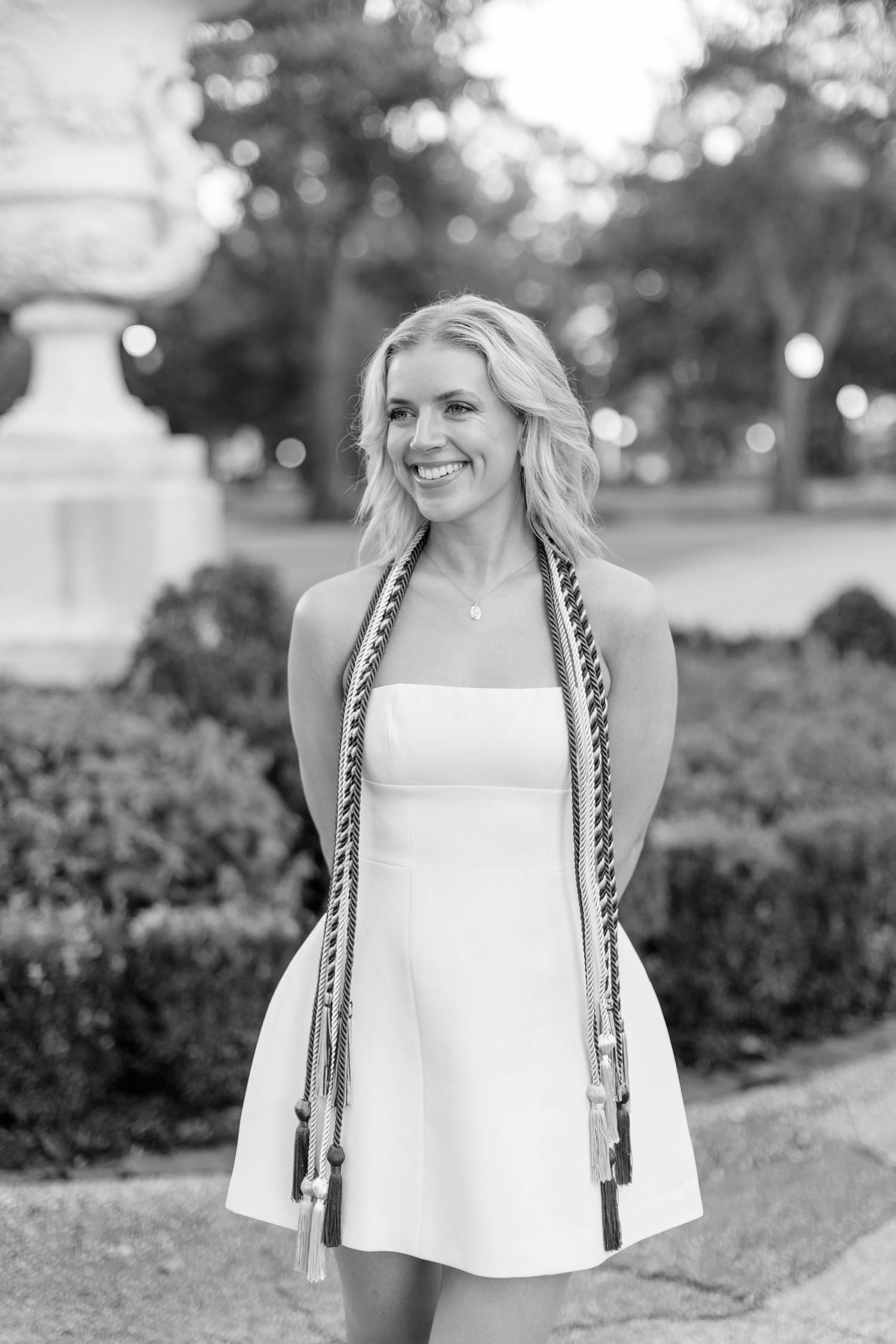 Black and white portrait captures graduation photo with decorative stone urn and foliage backdrop.