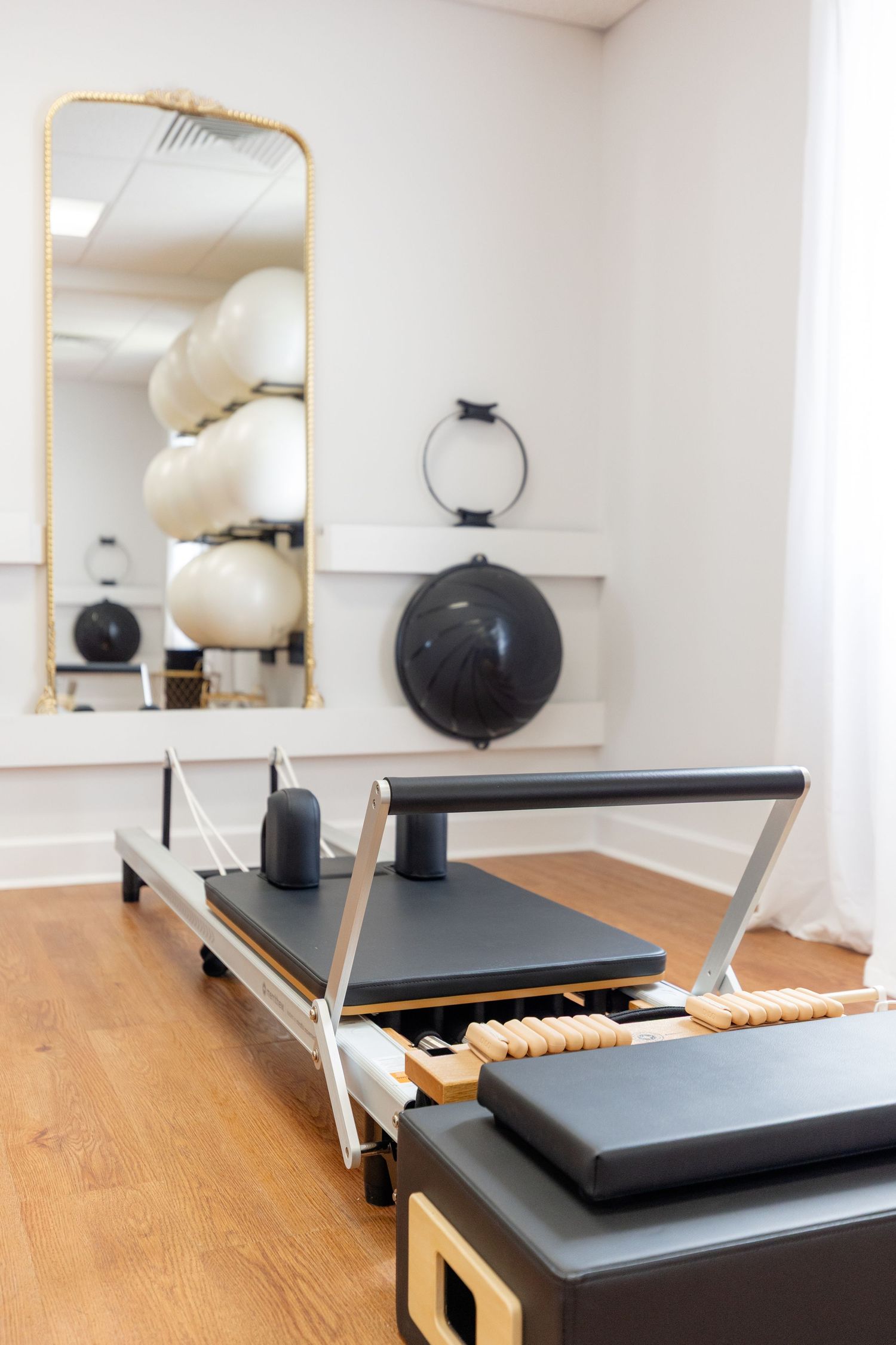 Pilates reformer machine with black cushions and wooden frame shown in a bright white studio with mirrors.