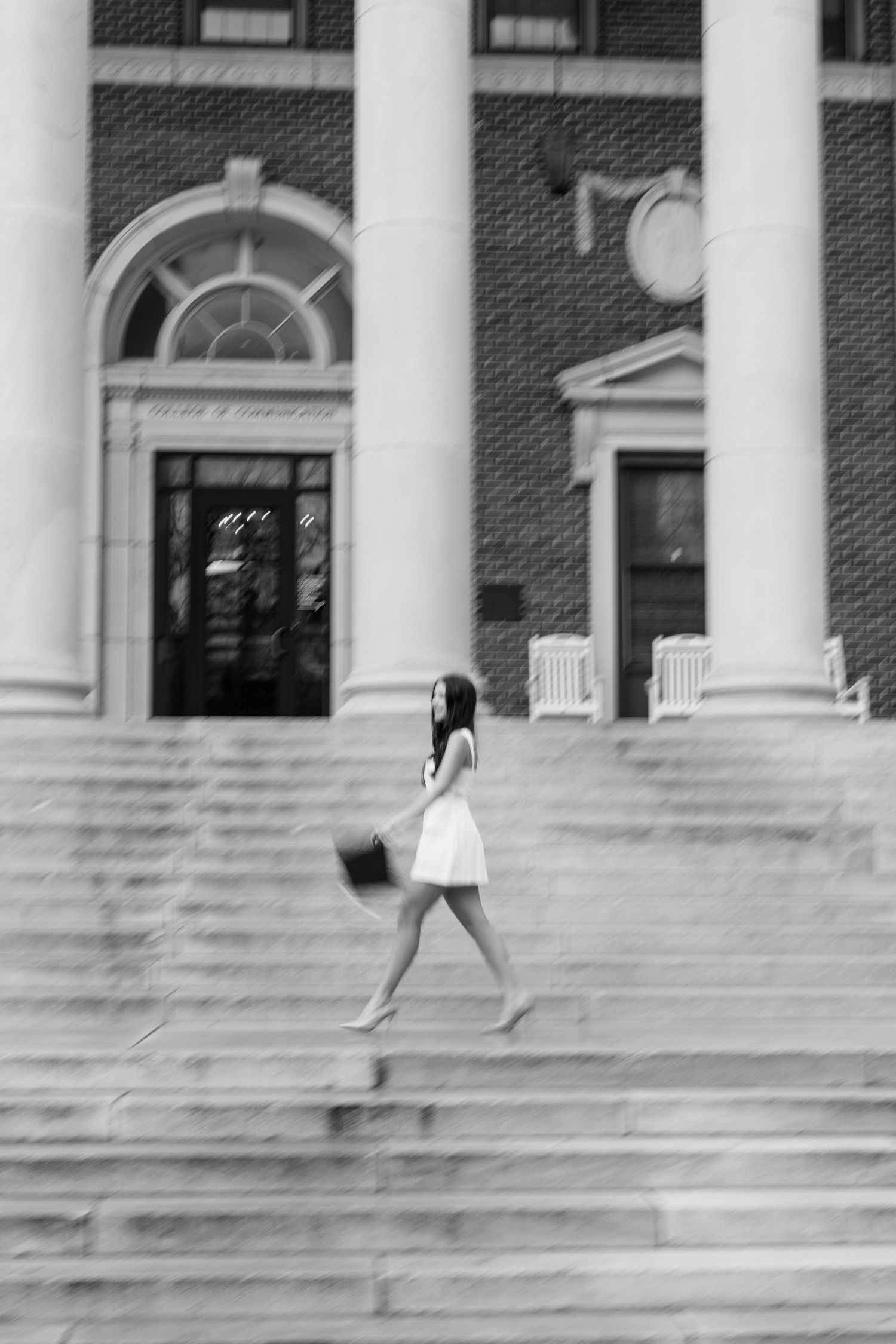 A person in a white dress walks down wide stone steps in front of a brick building with white columns.
