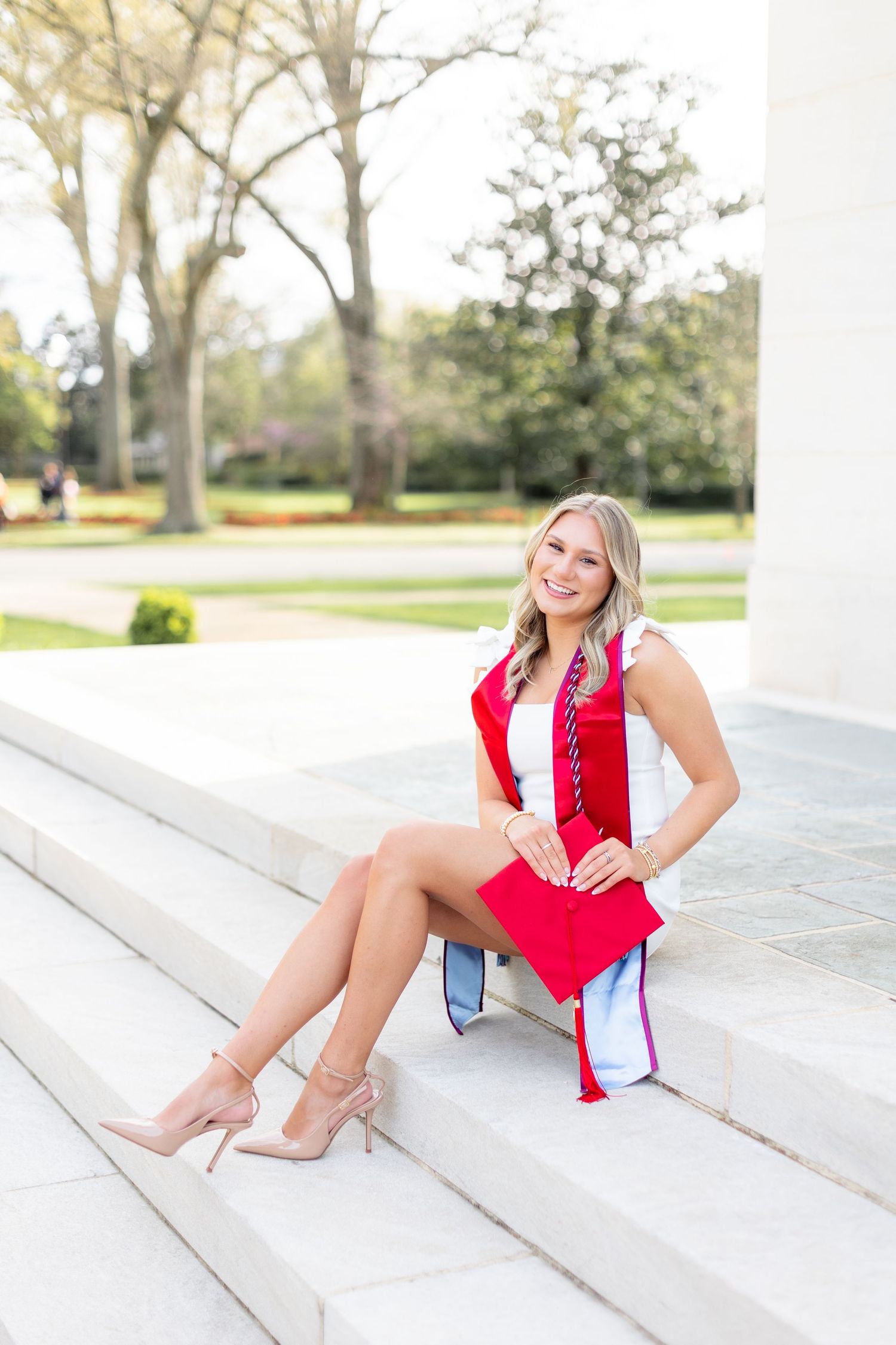 A person in a bright red dress sits gracefully on white stone steps outdoors on a sunny day.