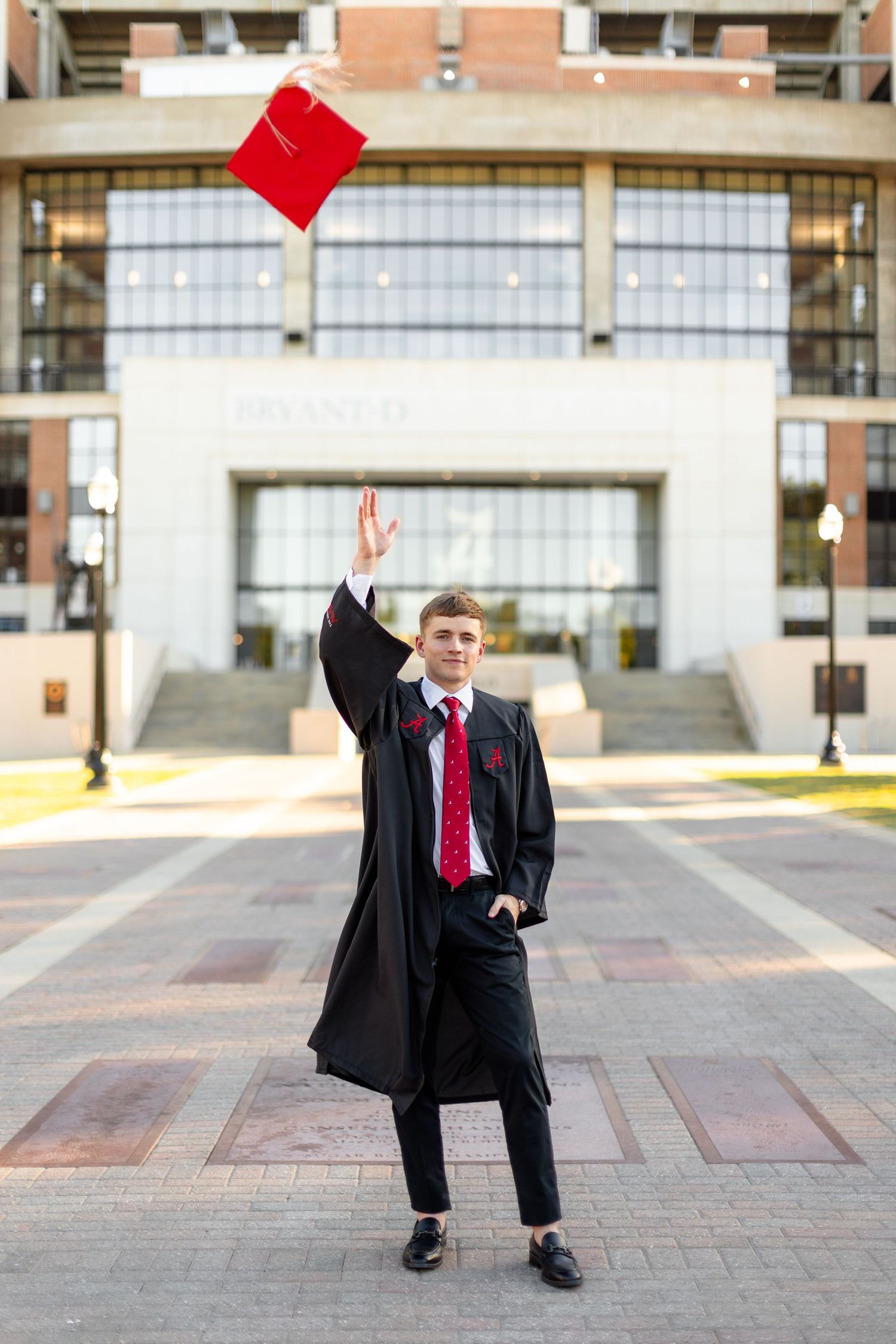 Graduate in black gown and red tie poses playfully with red graduation cap in outdoor courtyard setting.