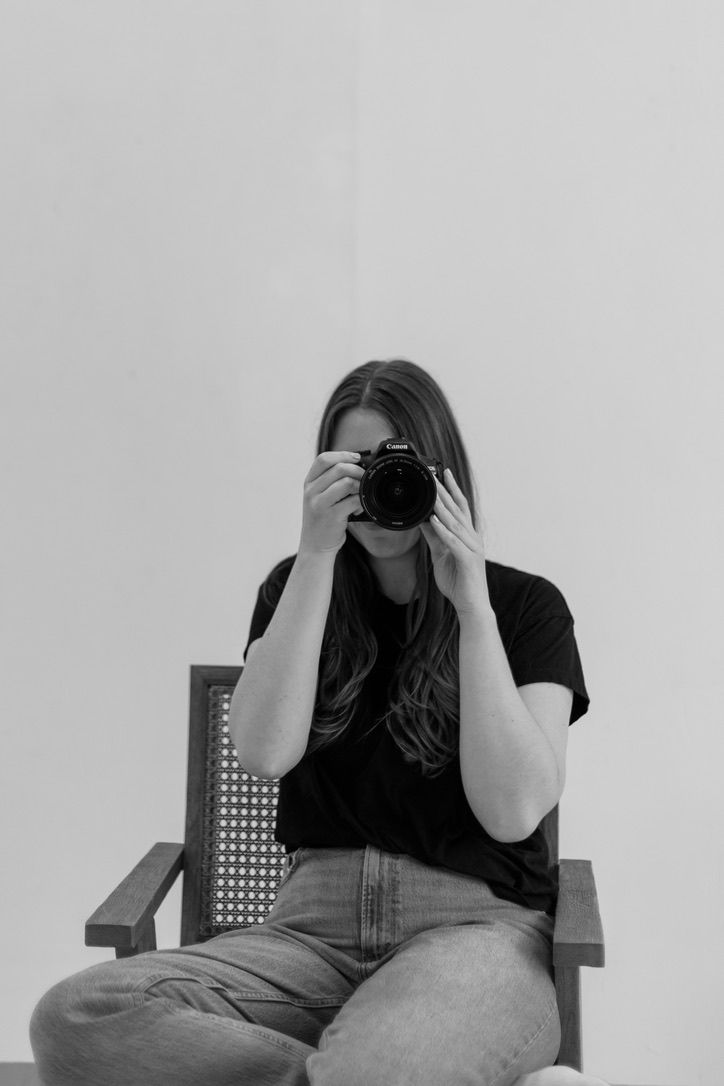 A photographer takes a self-portrait with a vintage camera while sitting in a wooden chair against a white wall.