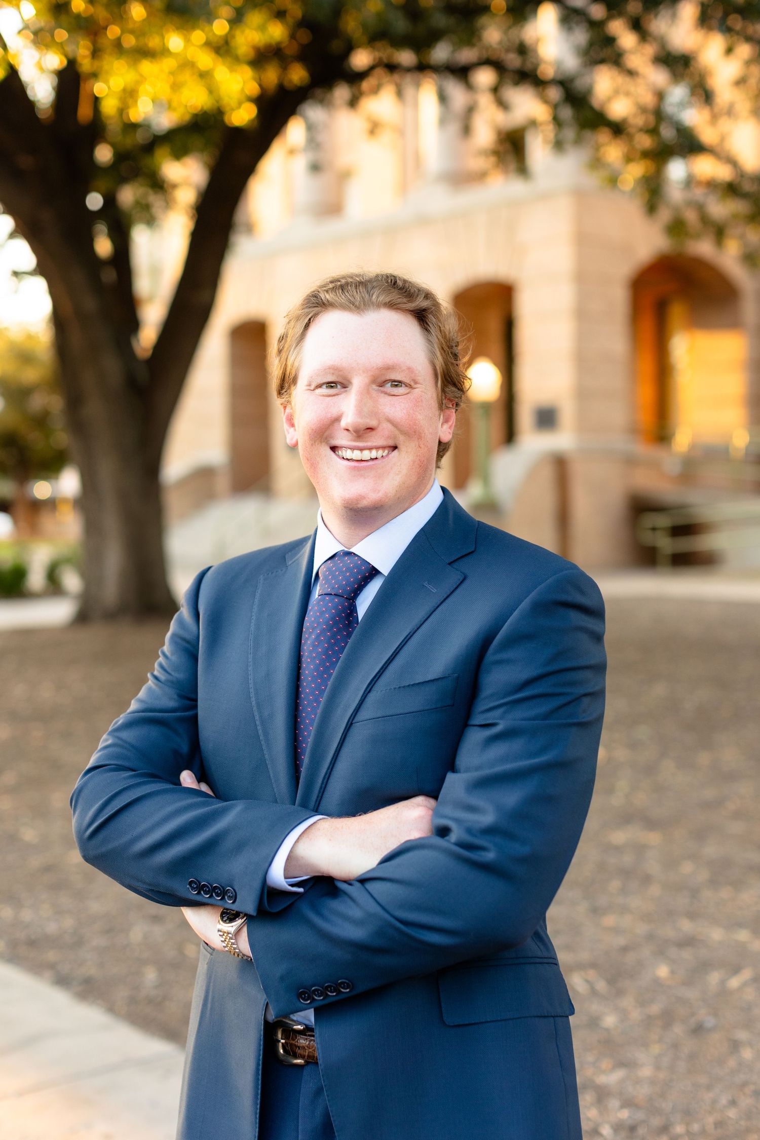 Professional portrait of someone in a navy suit and purple tie standing outdoors with autumn foliage and architecture visible.