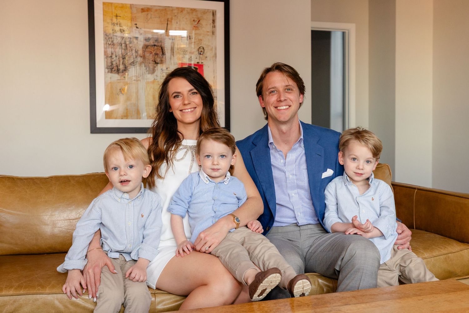 A family sits together on a beige couch in a bright living room with framed artwork on the walls.