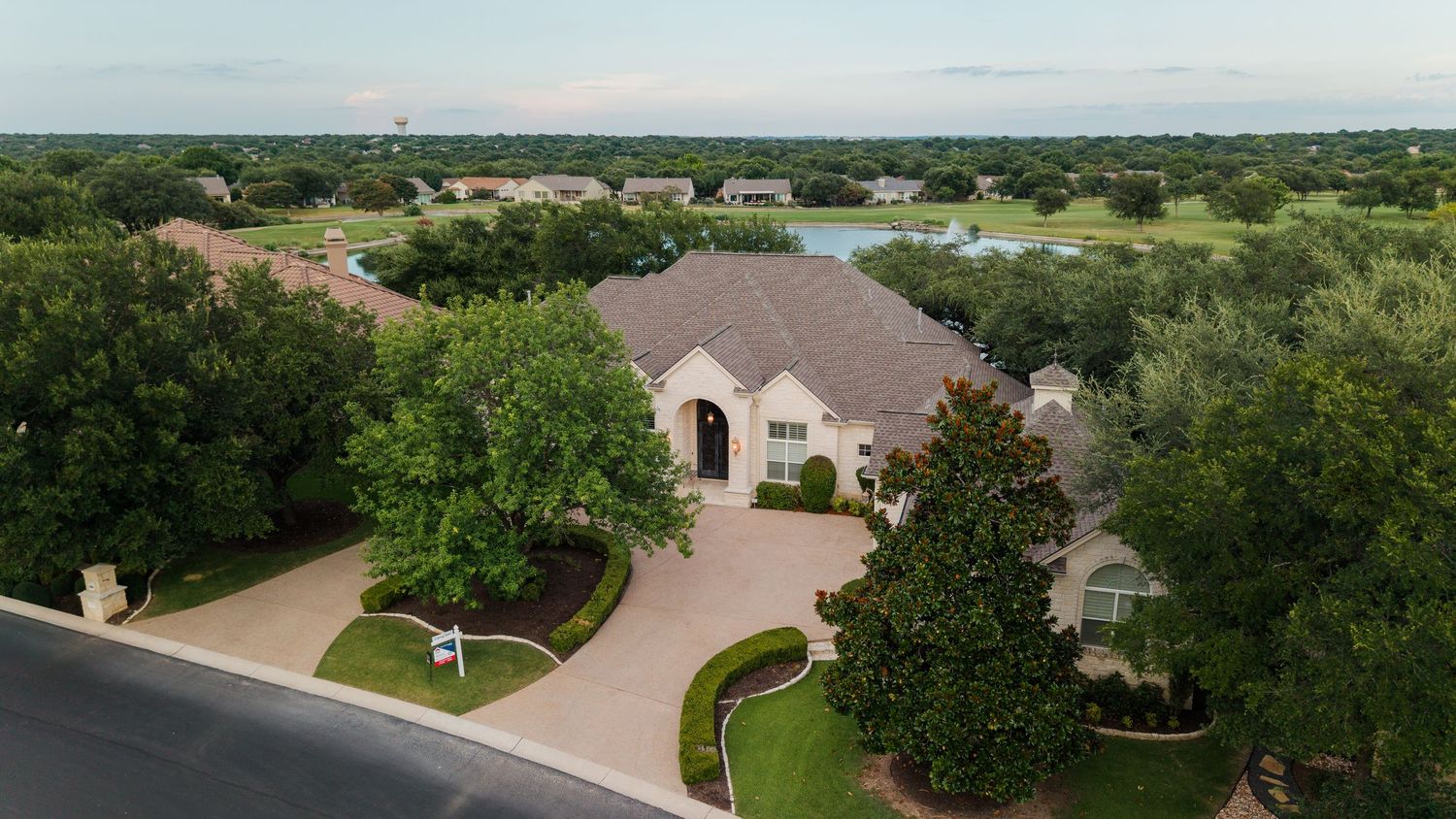 Aerial view of a luxury stone home with circular driveway, lush trees, and a serene lake in the background.