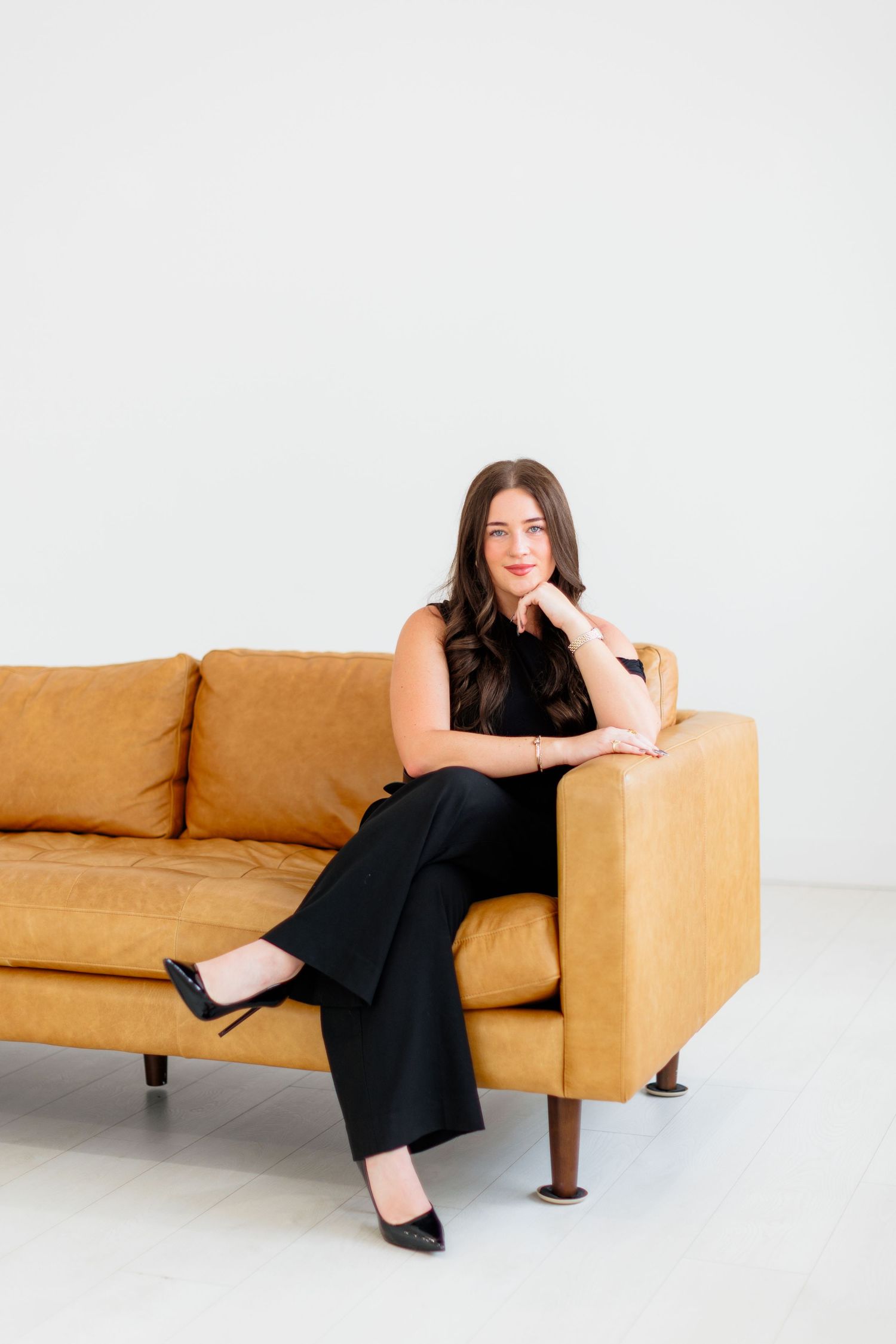 Professional woman in black outfit sitting confidently on a tan leather sofa against a white background.