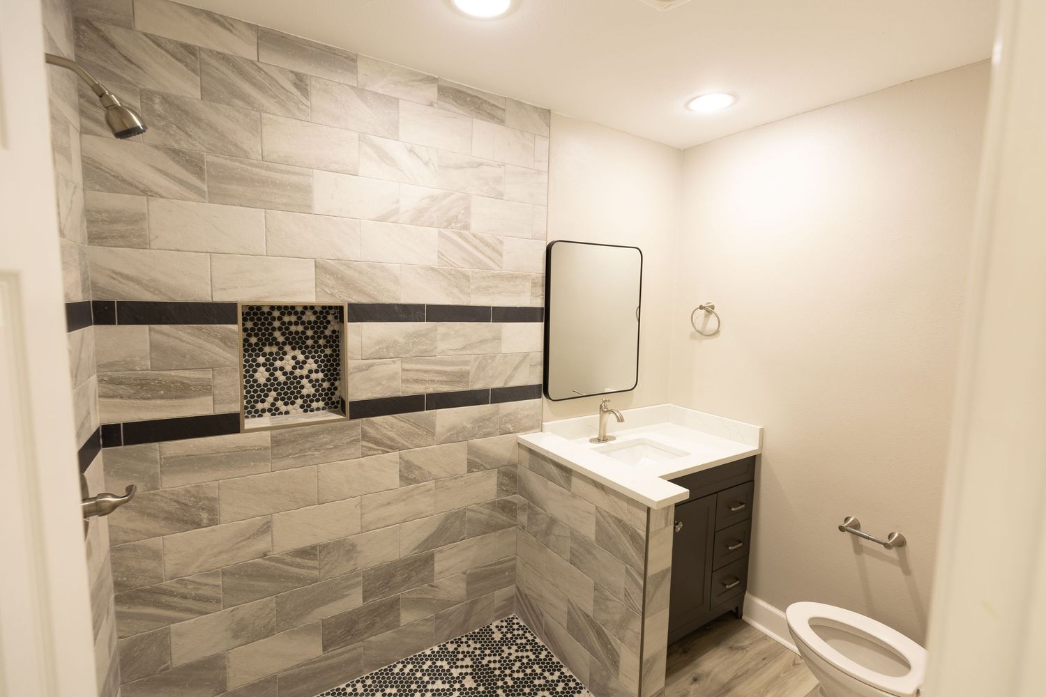 Modern bathroom with large gray tile shower, dark vanity, square mirror, and wood-look floors.