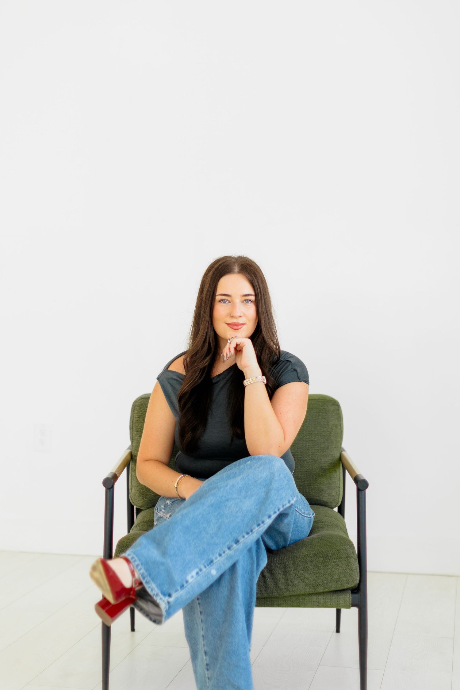 Brunette woman sitting in green chair wearing black top and jeans, posing confidently.