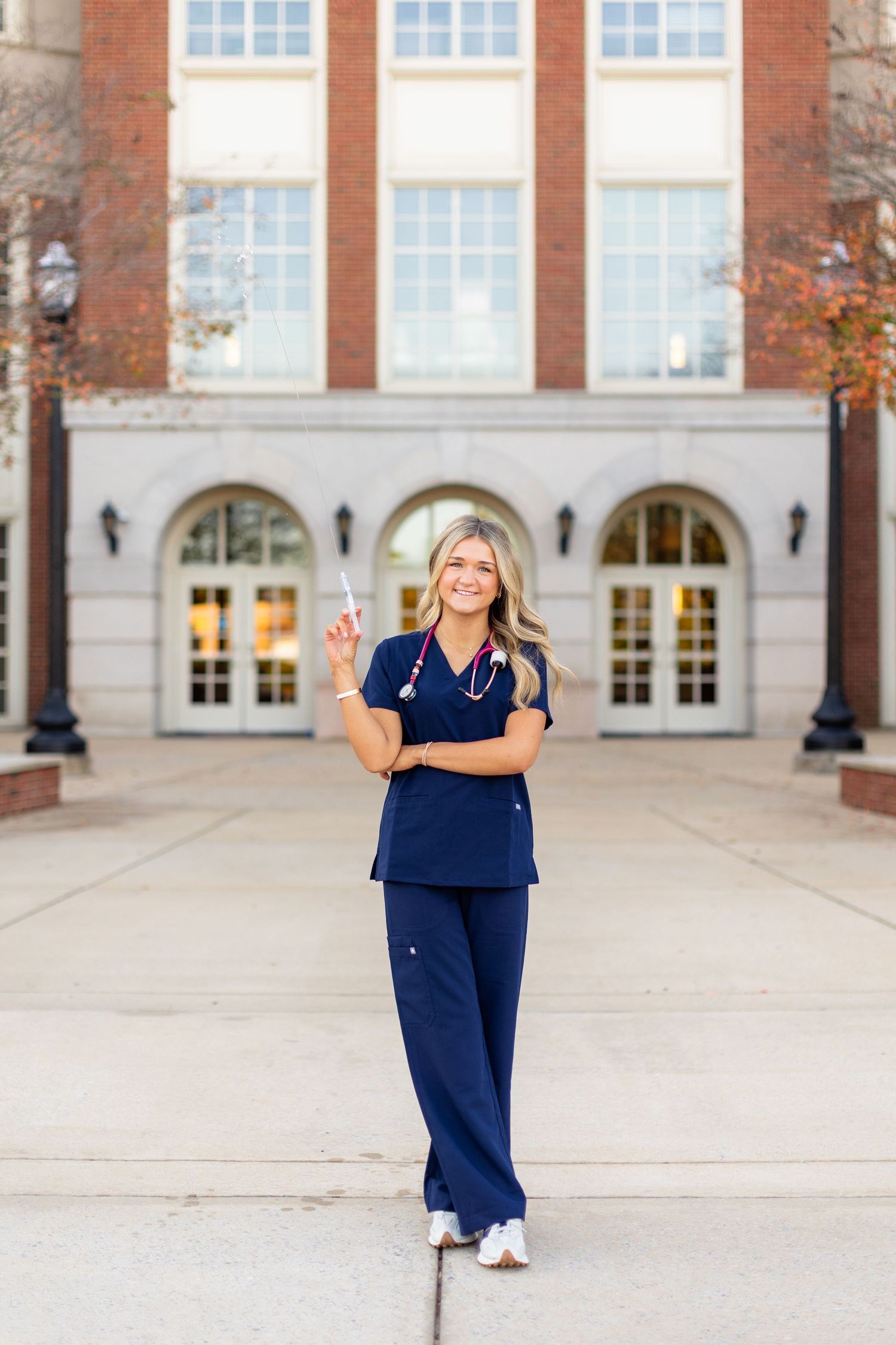 Healthcare professional in navy blue scrubs standing confidently in front of a brick building with arched windows.