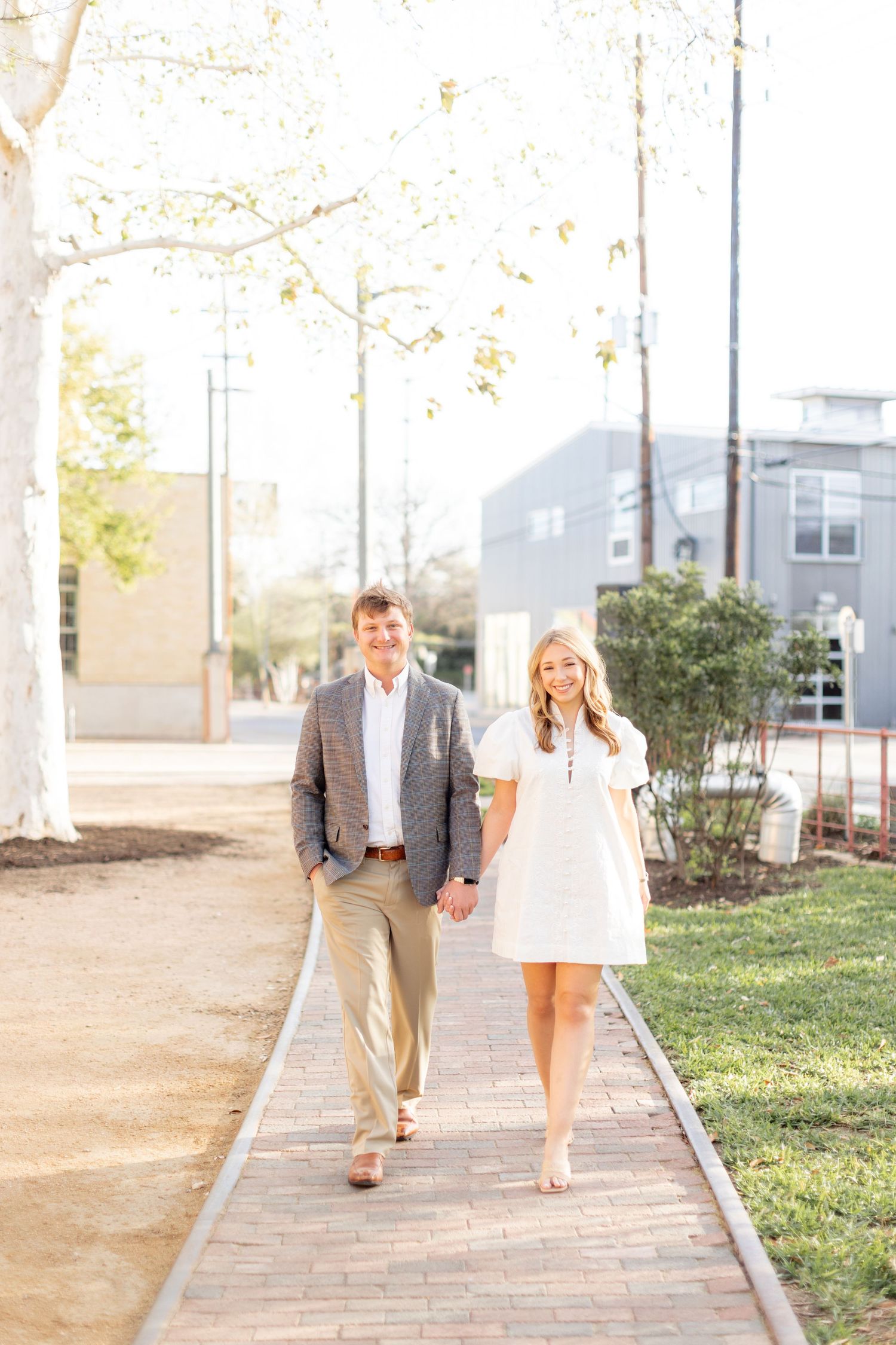 A couple walks together on a paved pathway through a sunny outdoor garden setting.
