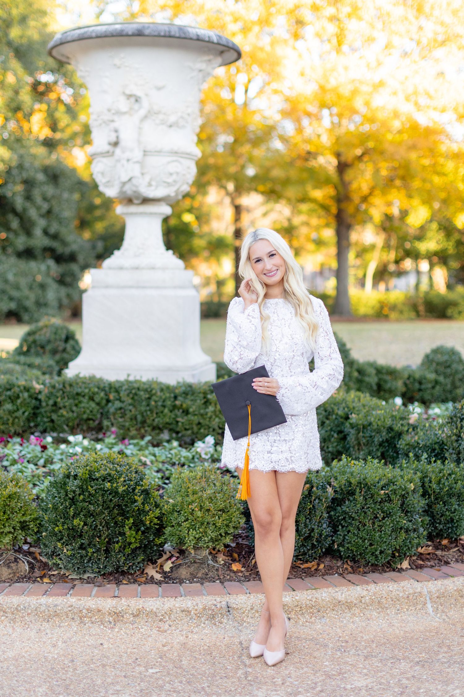 Classic white stone urn and garden pathway backdrop frames a graduation portrait in elegant white lace dress.