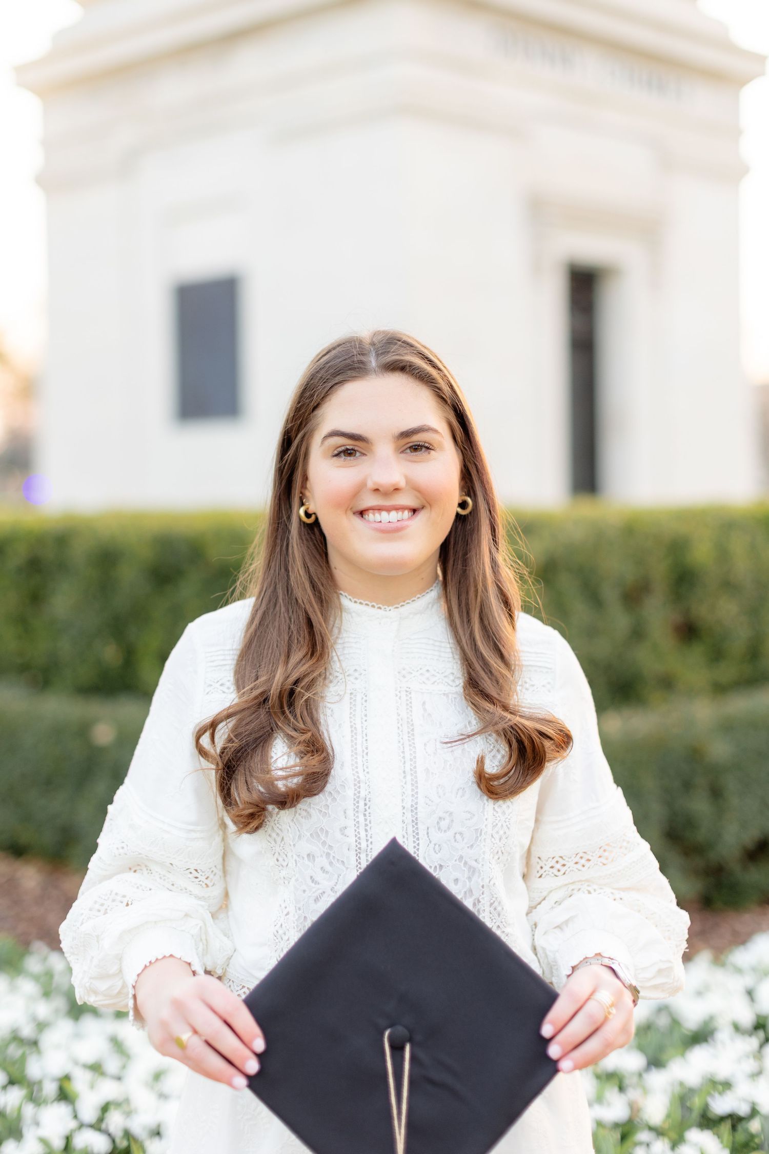 A graduate in a white knit sweater holds a black graduation cap while standing in front of white flowers and greenery.