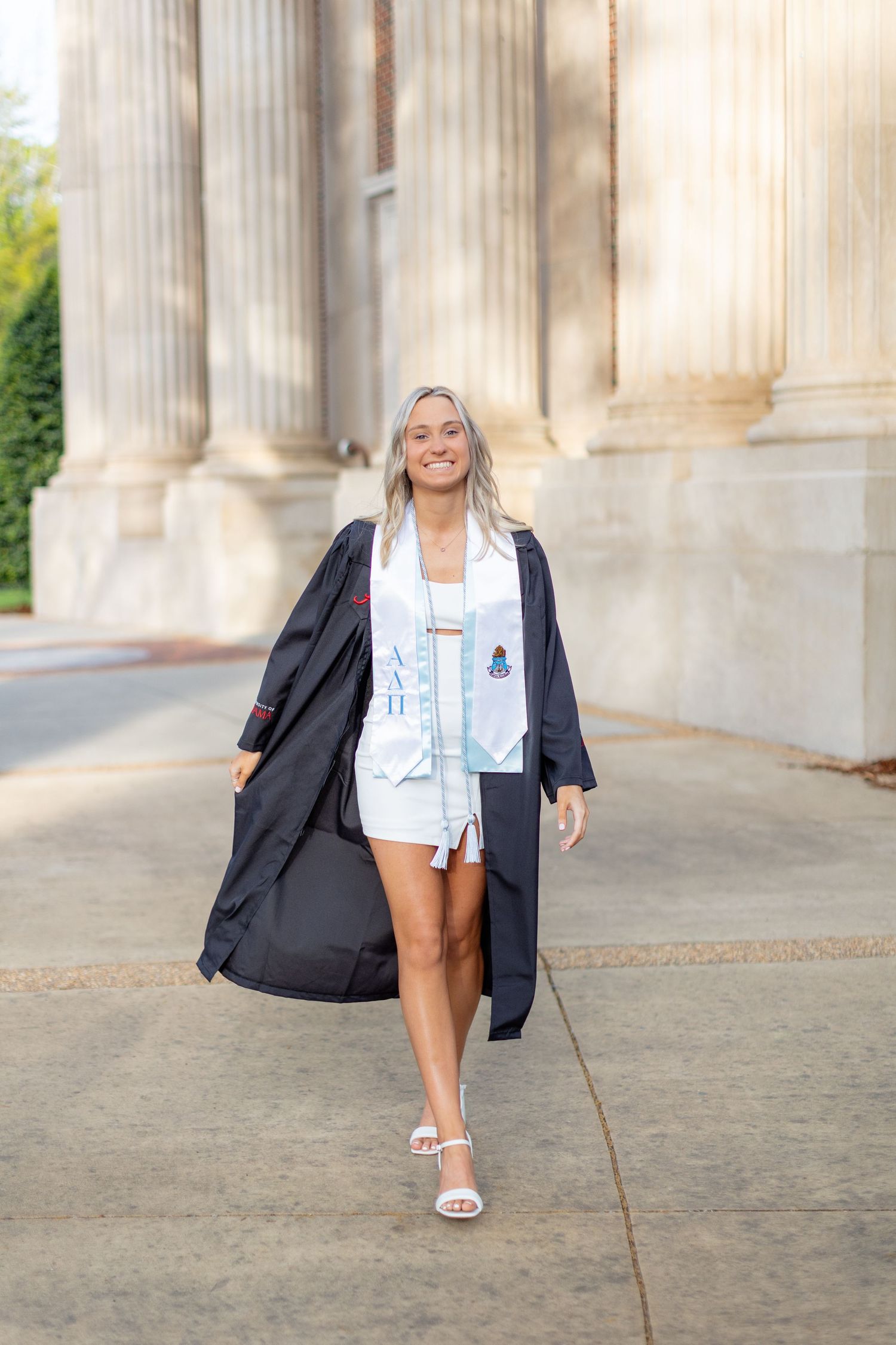 A graduate in cap and gown walking confidently past classic stone columns in summer.