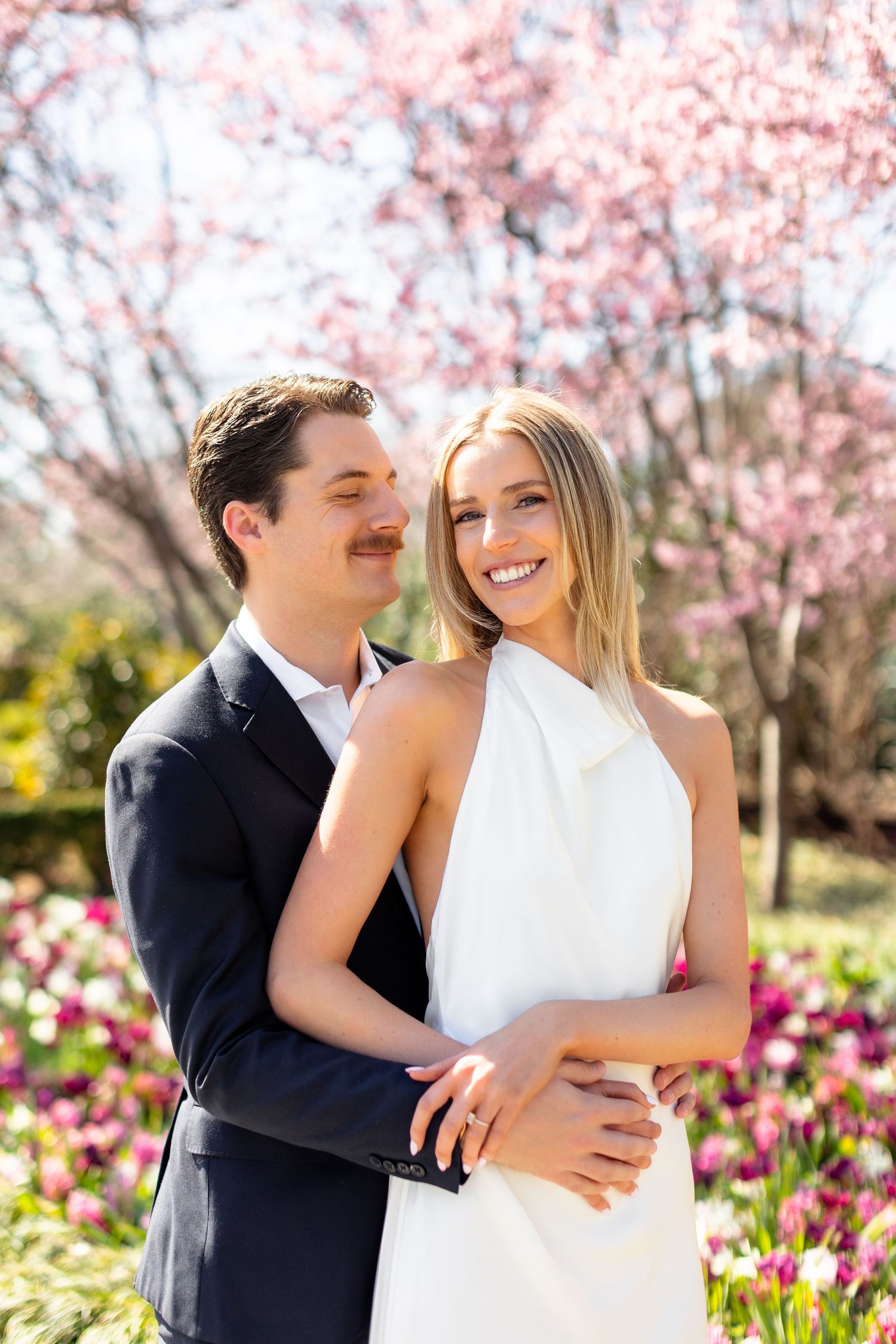 A couple embraces in a garden with blooming cherry blossoms and pink tulips on a sunny spring day.