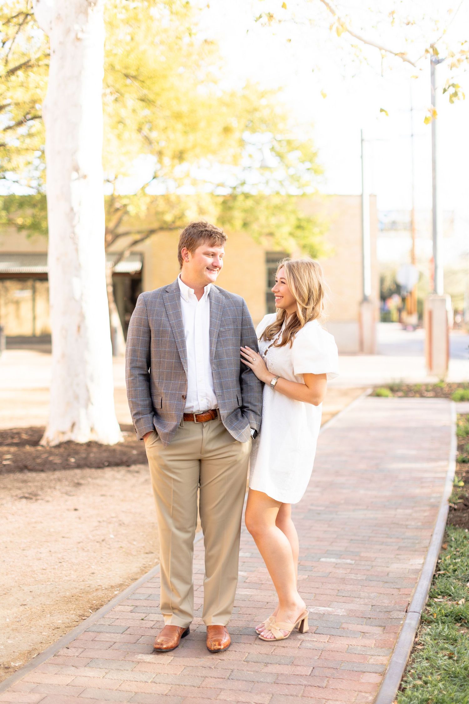A couple stands together on a brick path, with the person in a white dress smiling at their partner in a gray blazer.