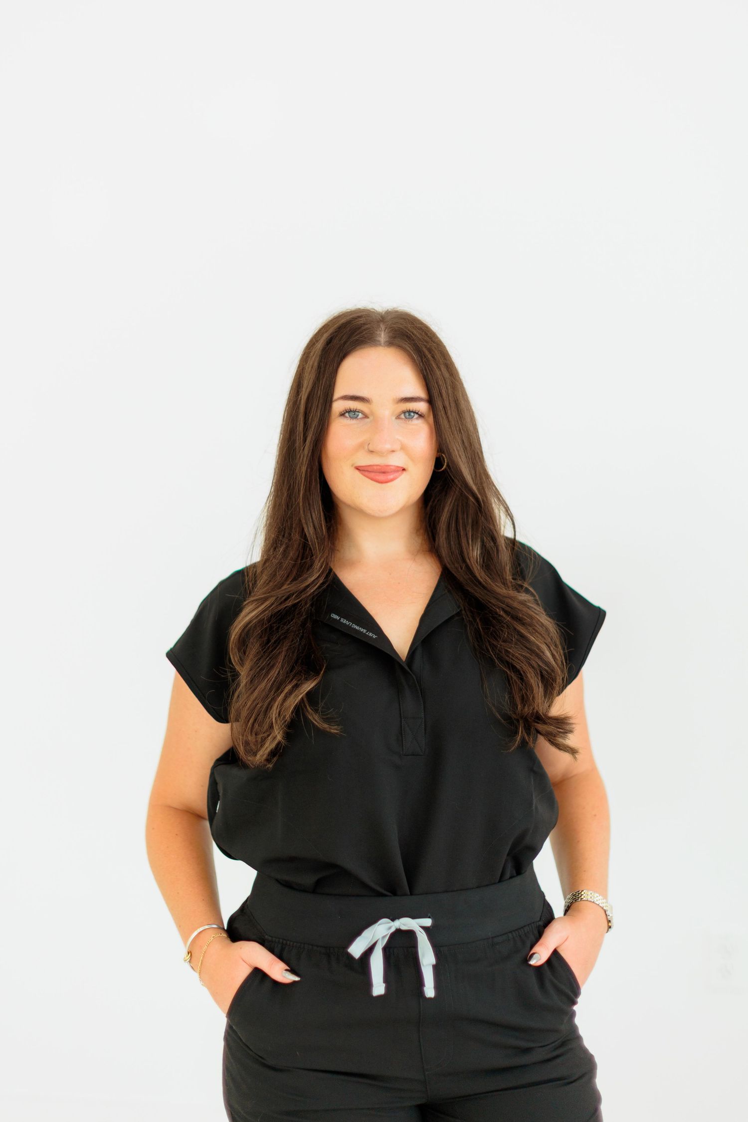 Smiling brunette woman in black jumpsuit standing against white background with hands in pockets.