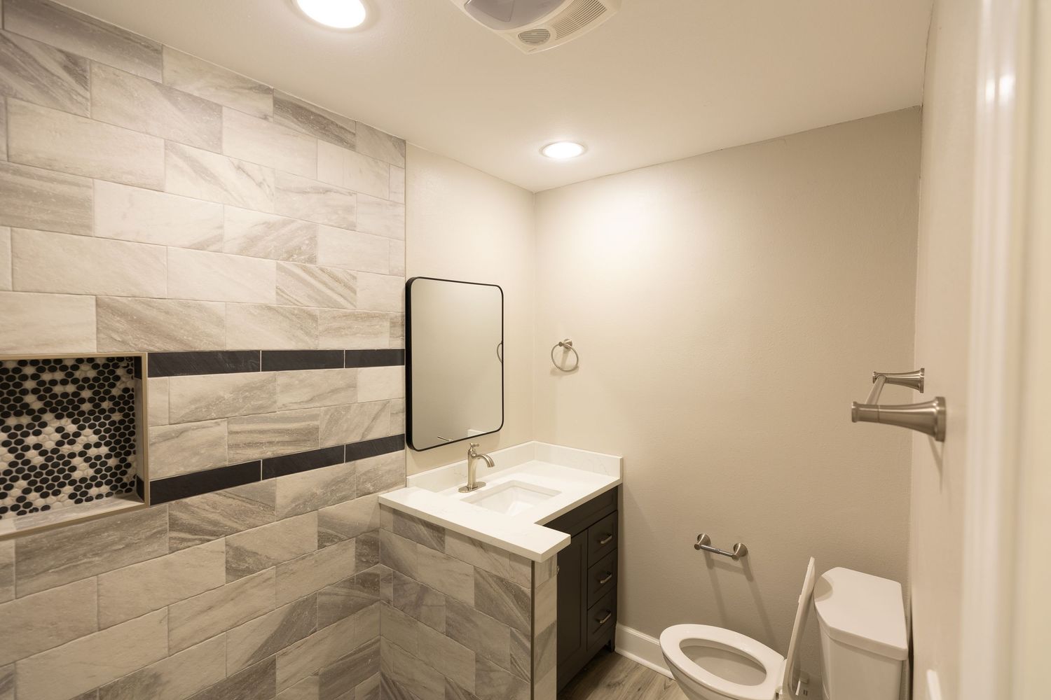 Renovated bathroom featuring gray tile accent wall, white vanity, round mirror, and toilet.