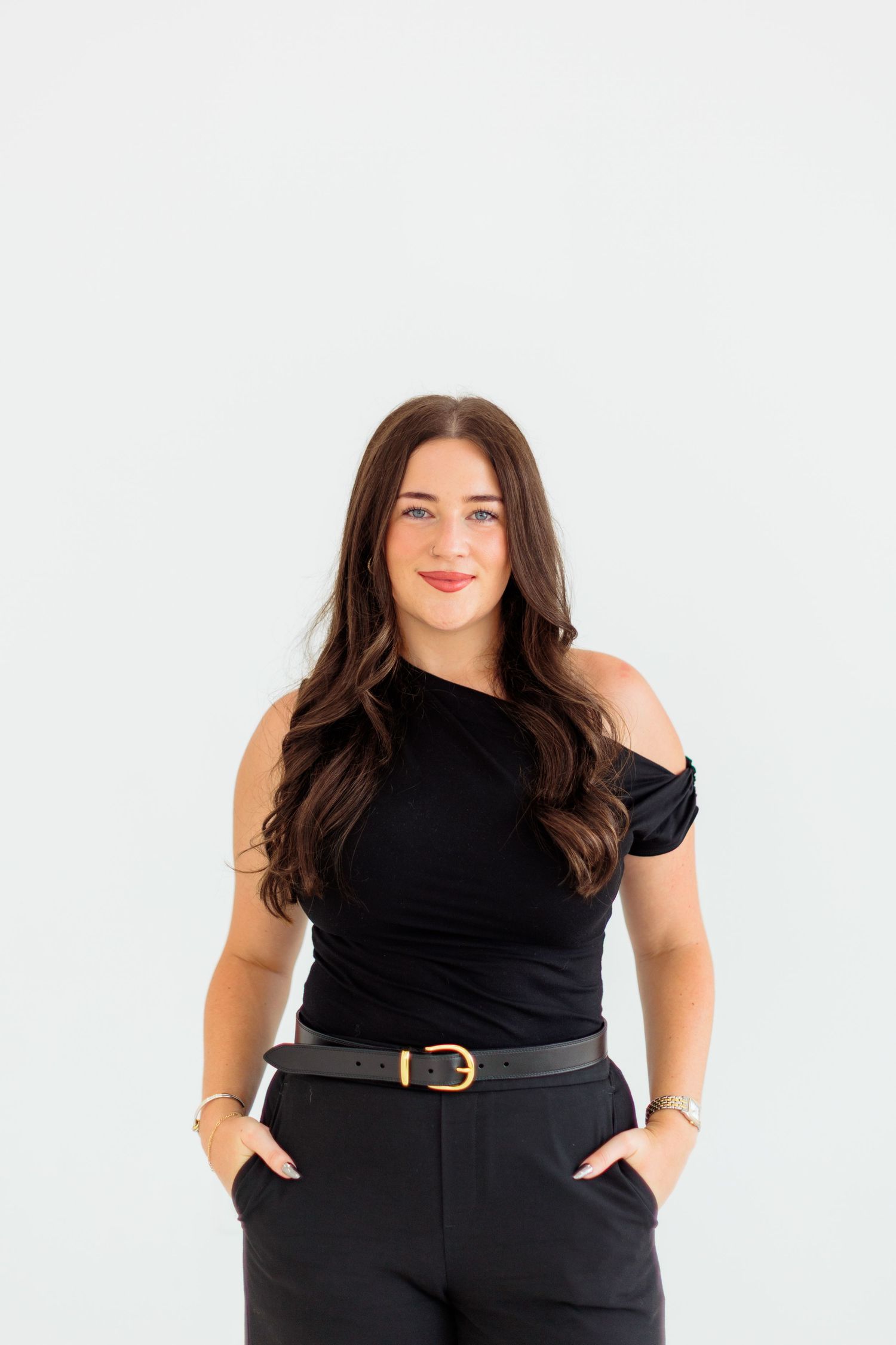 Brunette woman in black one-shoulder top and belt smiling confidently.