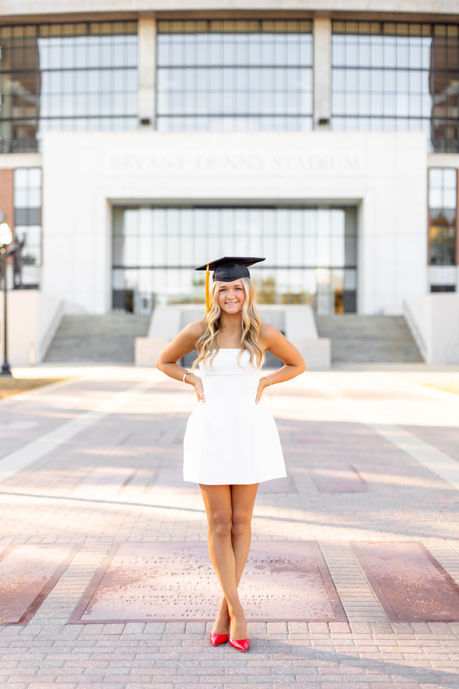Graduate in white dress and cap poses on brick courtyard with modern building in background.