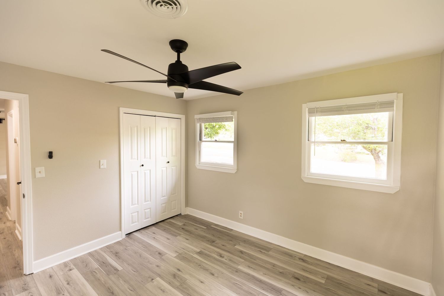 Bright empty bedroom with light wood floors, white closet door, two windows, and a modern black ceiling fan.