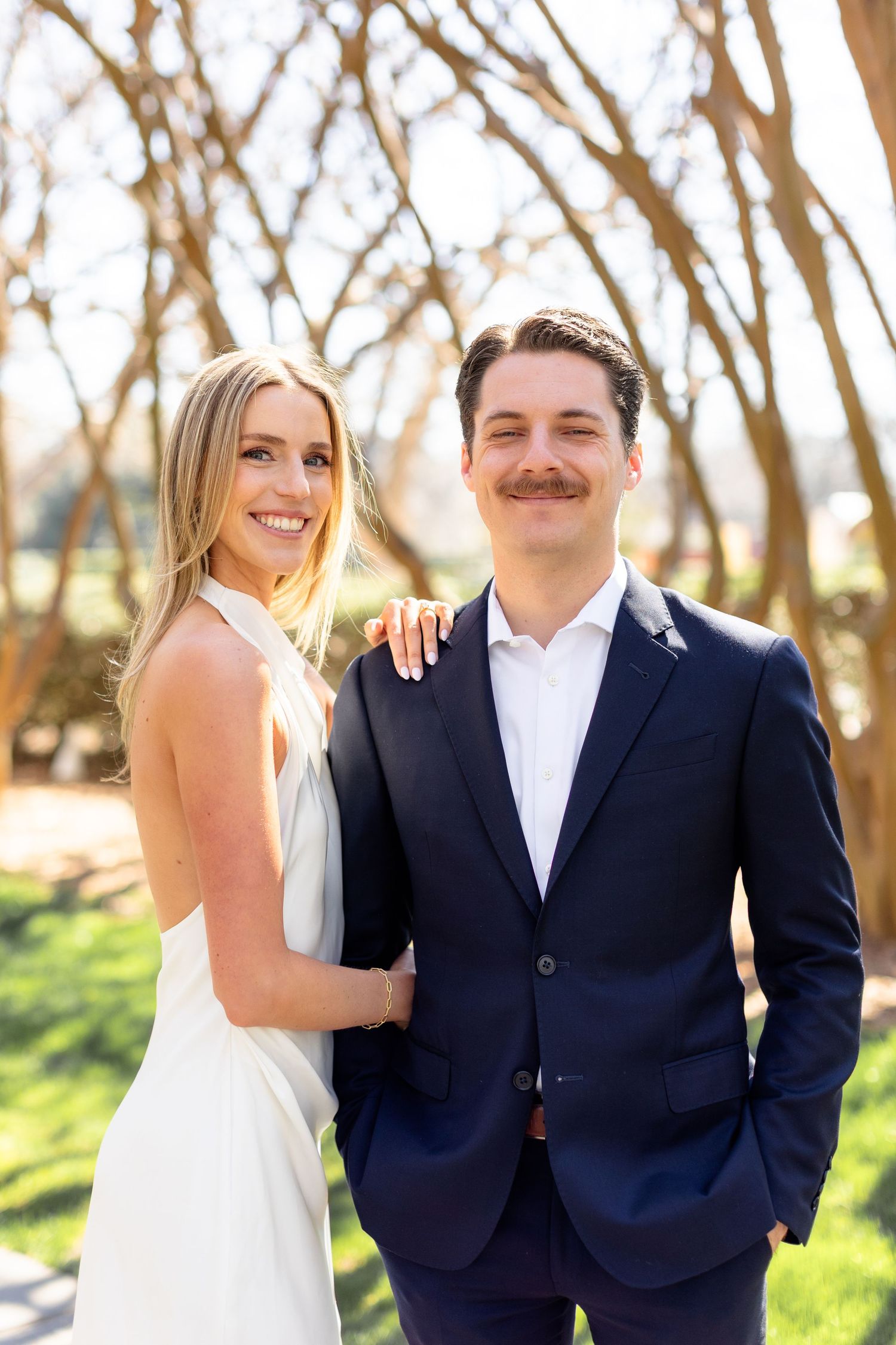 A couple in wedding attire smiles together in a sunny outdoor setting with trees in the background.