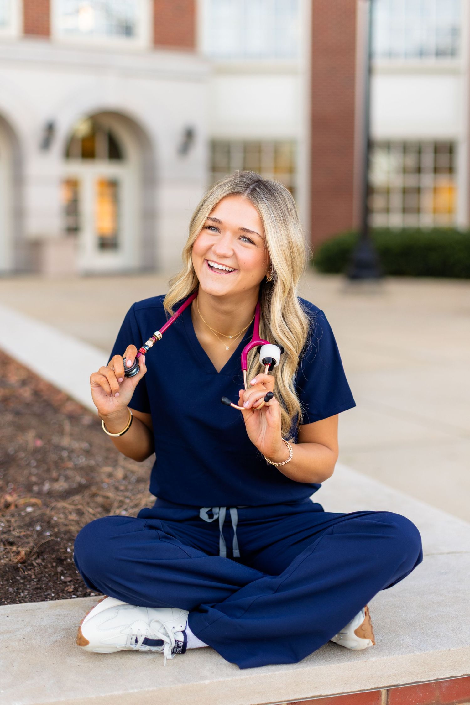 Healthcare worker in navy scrubs sits cross-legged on ground while holding medical supplies and smiling warmly.