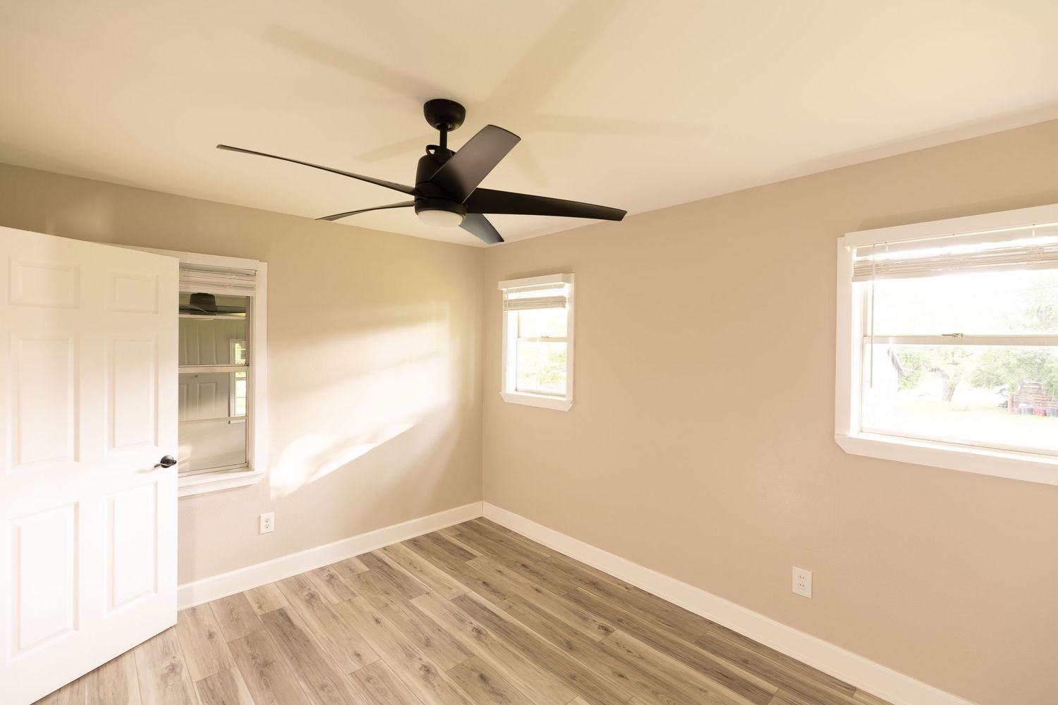 Sunlit empty bedroom with hardwood floors, windows, and a black ceiling fan.