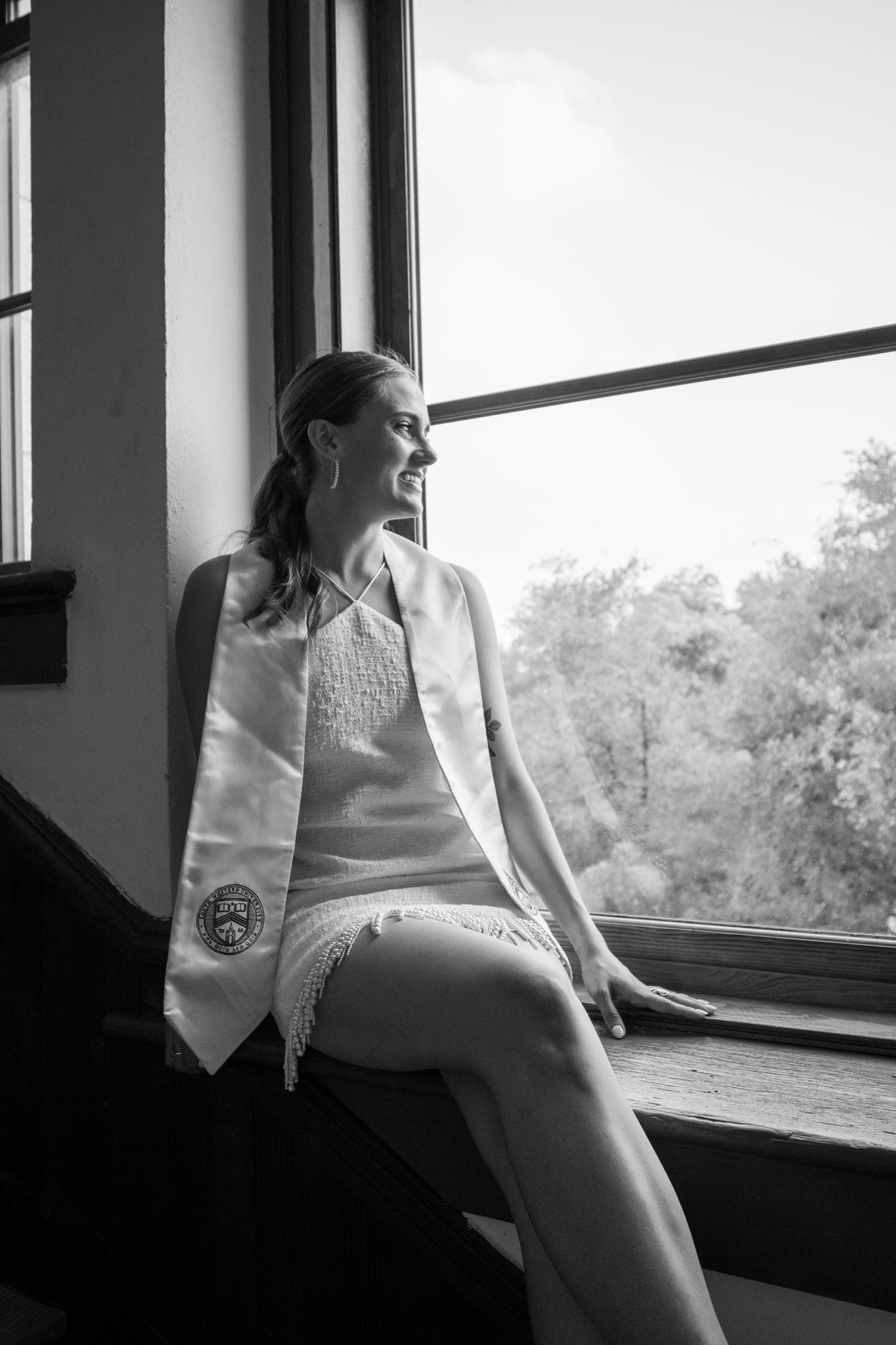 A black and white photo of someone in graduation attire sitting contemplatively by a window.