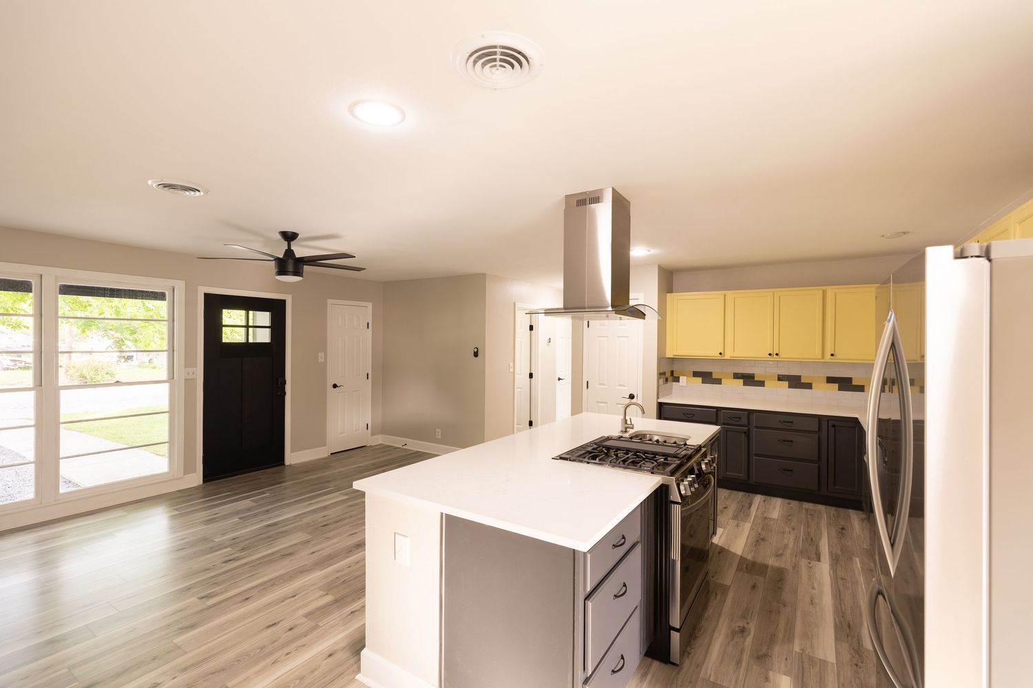 Modern kitchen with yellow upper cabinets, island with cooktop, stainless steel hood vent, and open living area with hardwood floors.