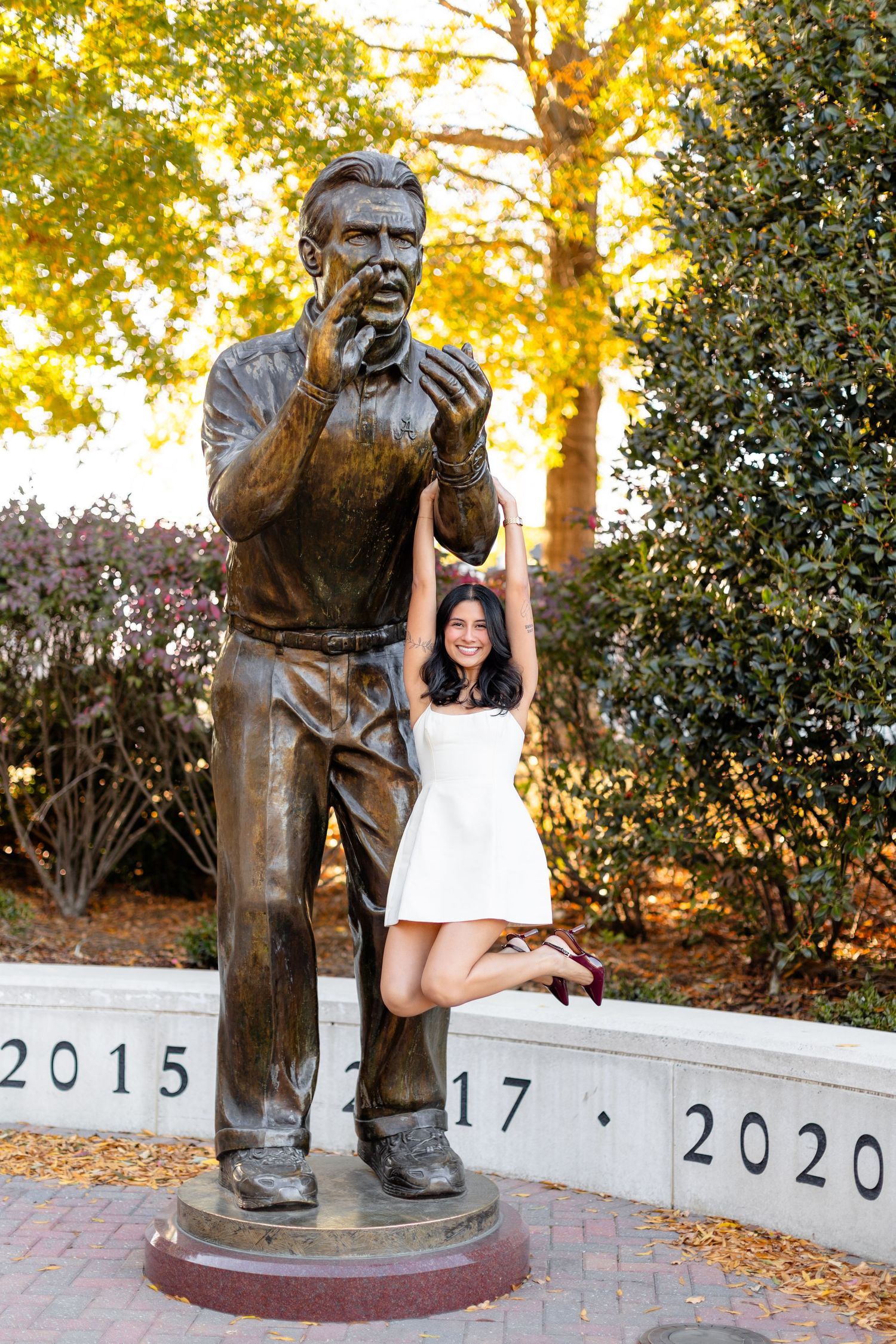 A bronze statue stands on a memorial pedestal while someone in a white dress poses playfully next to it at sunset.