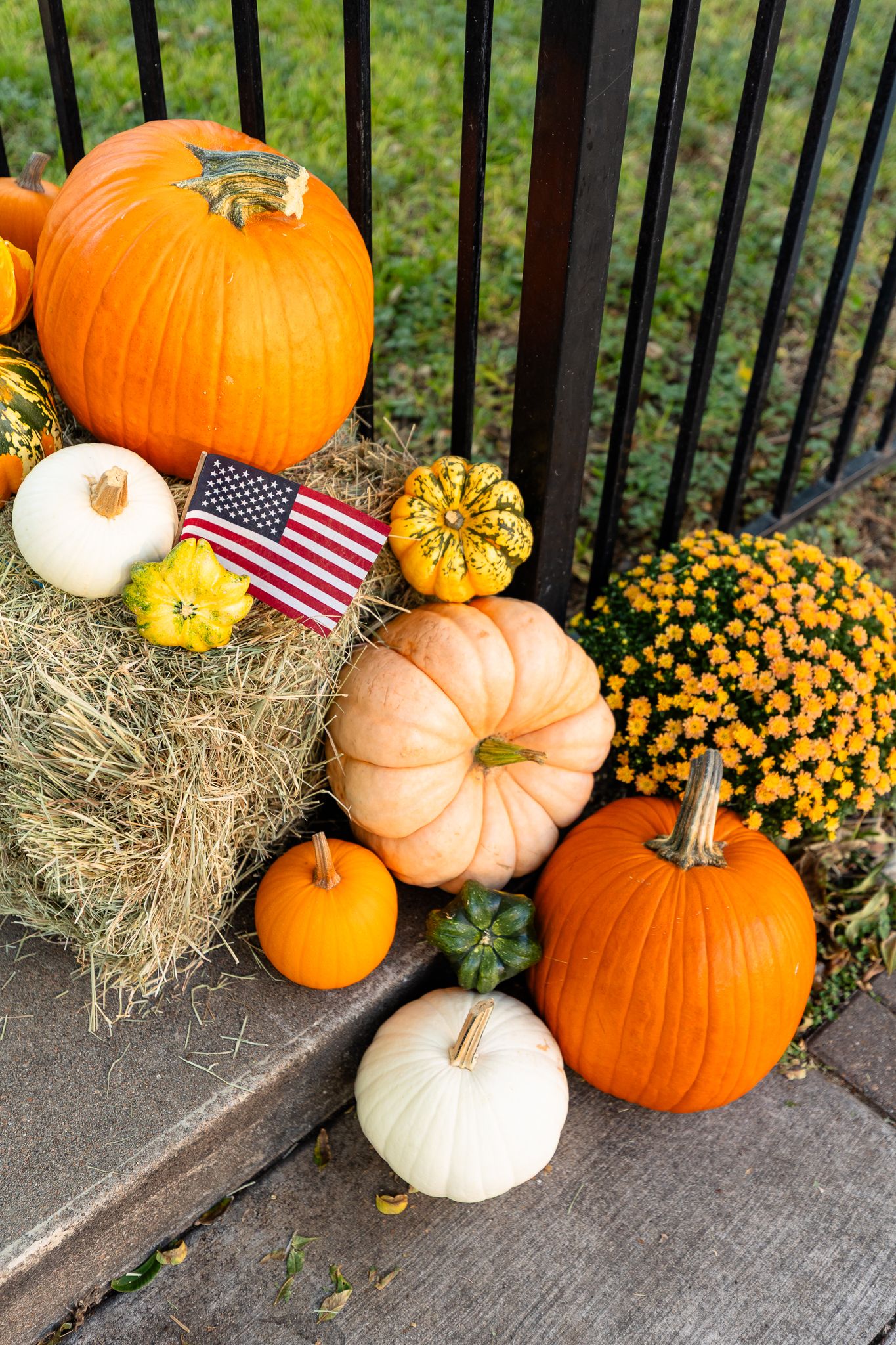 Fall harvest display with orange and white pumpkins, yellow mums, hay bales and small American flag on porch steps.