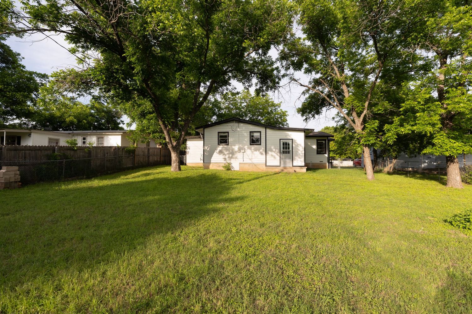 White ranch home with large mature trees, green lawn, and wood privacy fence in backyard.