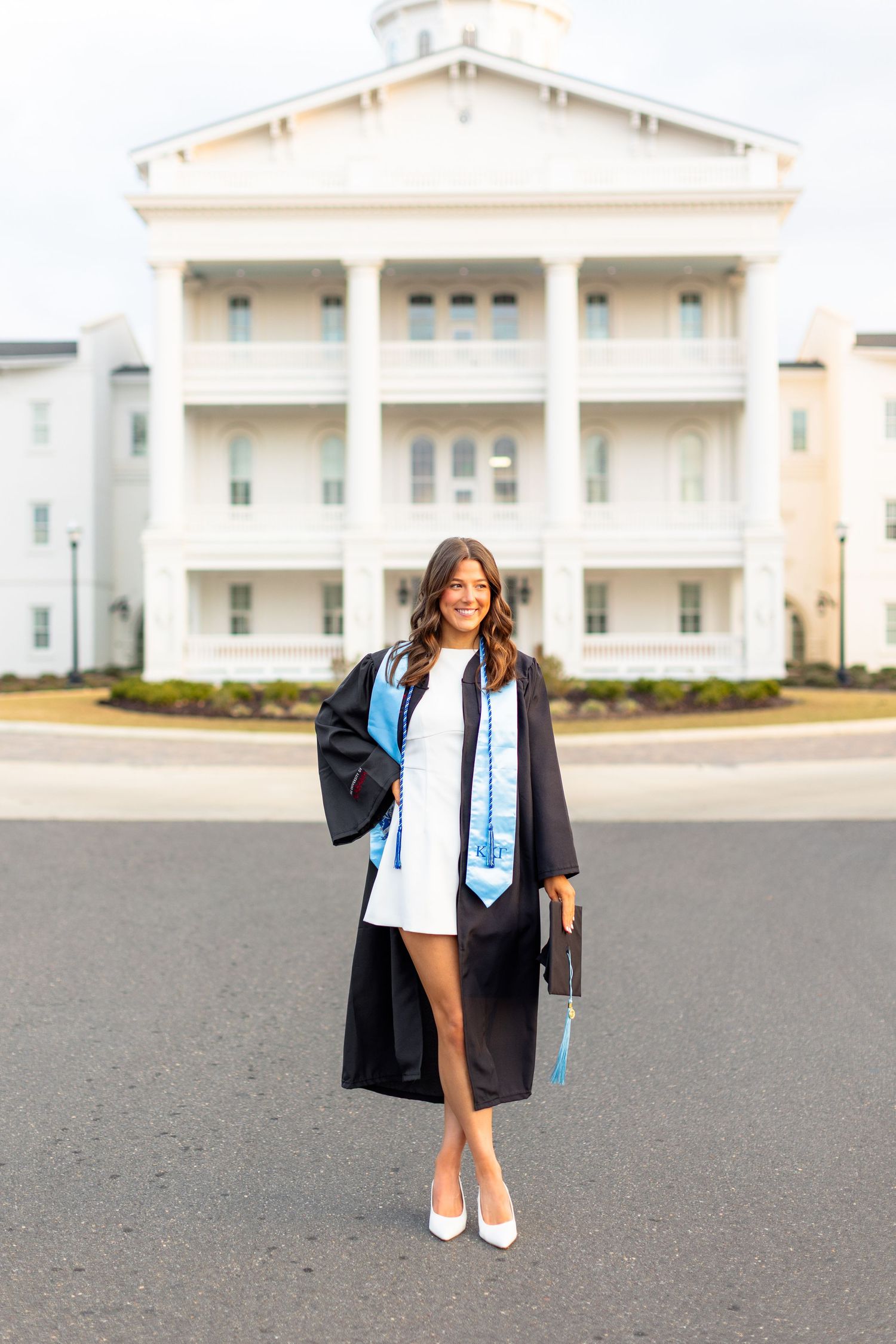 Graduate in cap and gown poses in front of white university building during commencement celebration.
