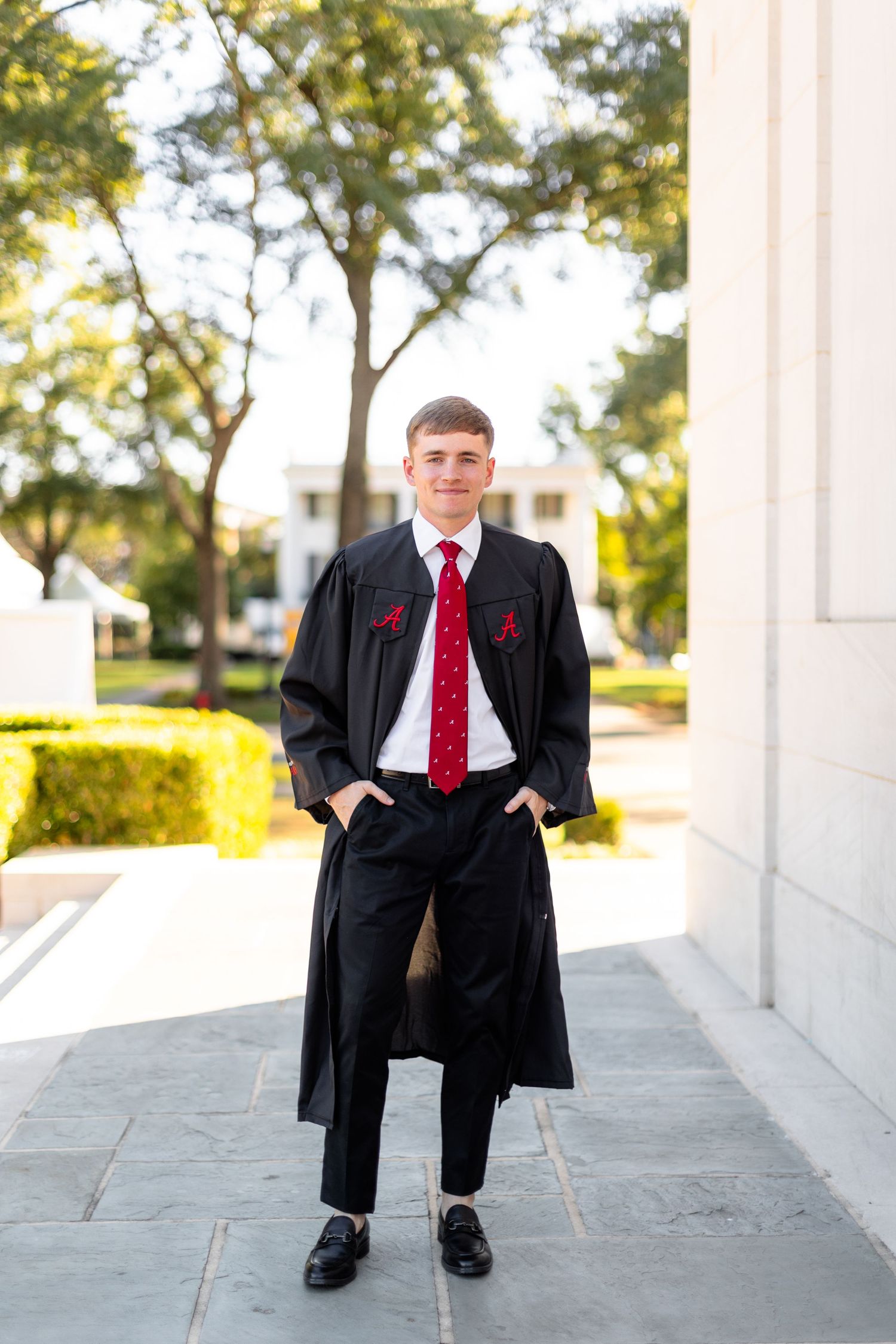 Graduate in black academic regalia and red tie stands on stone porch with trees and sunlight in background.