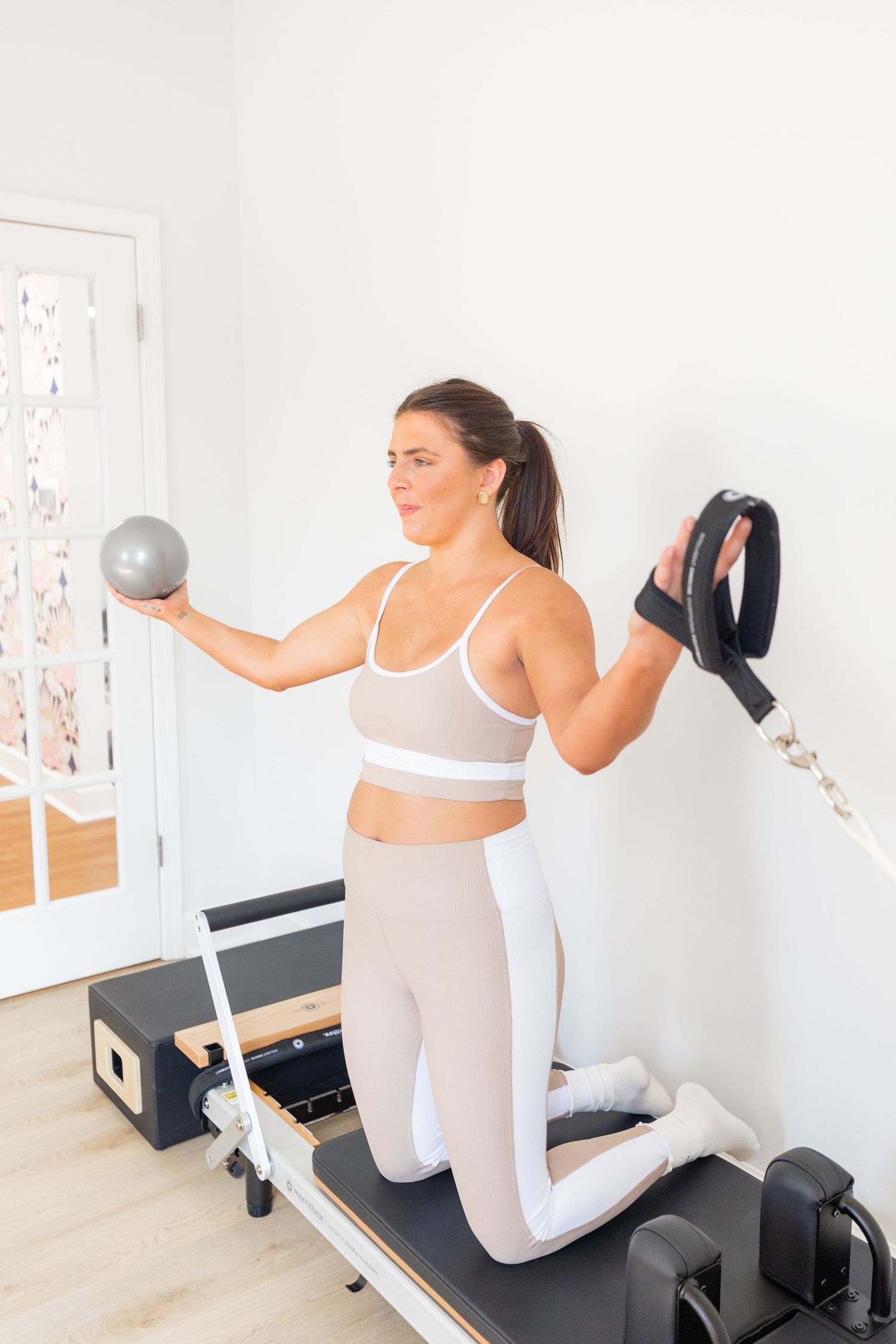 Person in white athletic wear demonstrates proper form for Pilates reformer exercises in a bright studio space.