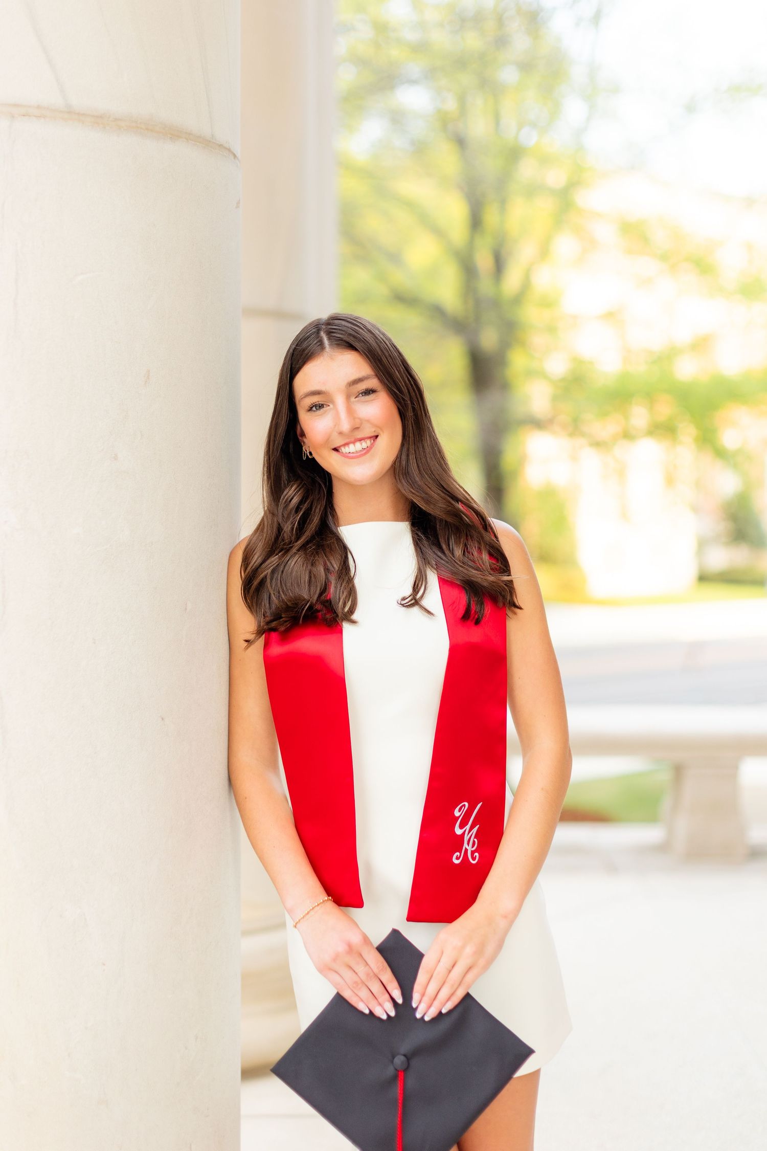 A graduate in academic regalia poses against white columns outdoors wearing a red stole and black gown.