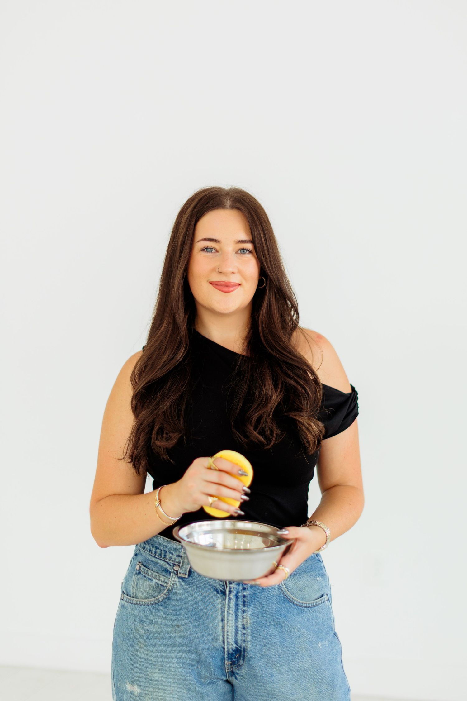 Young woman in black top and jeans holding a lemon and silver mixing bowl against white background.