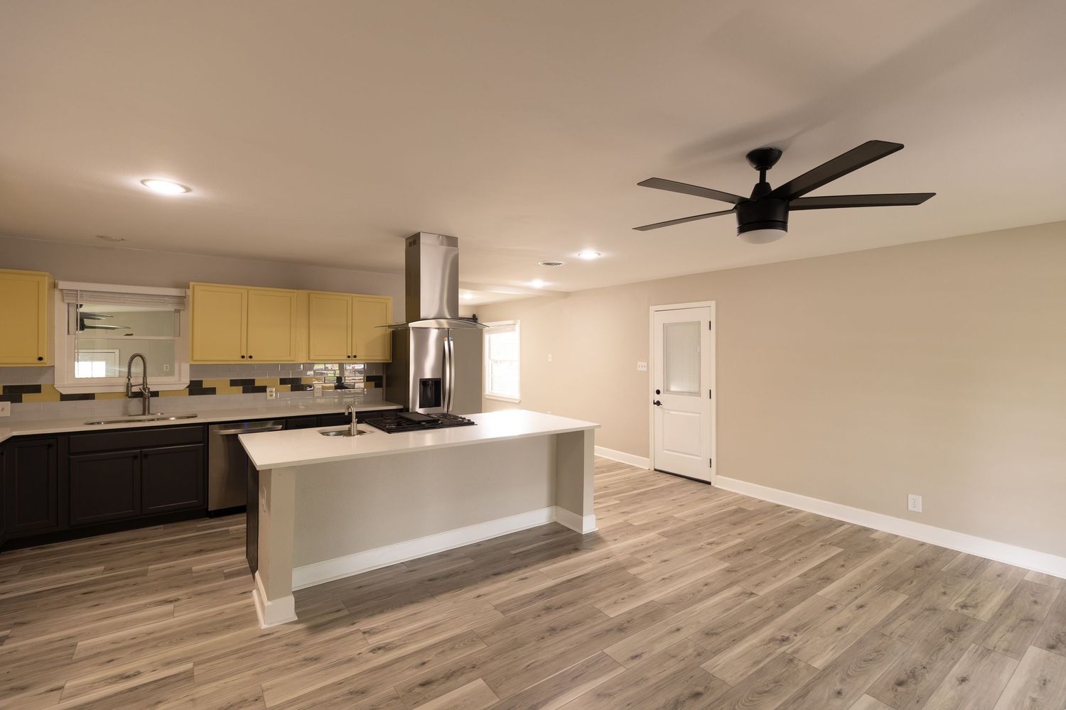 Modern open-concept kitchen with island, ceiling fan, hardwood floors, yellow cabinets, and stainless steel range hood.