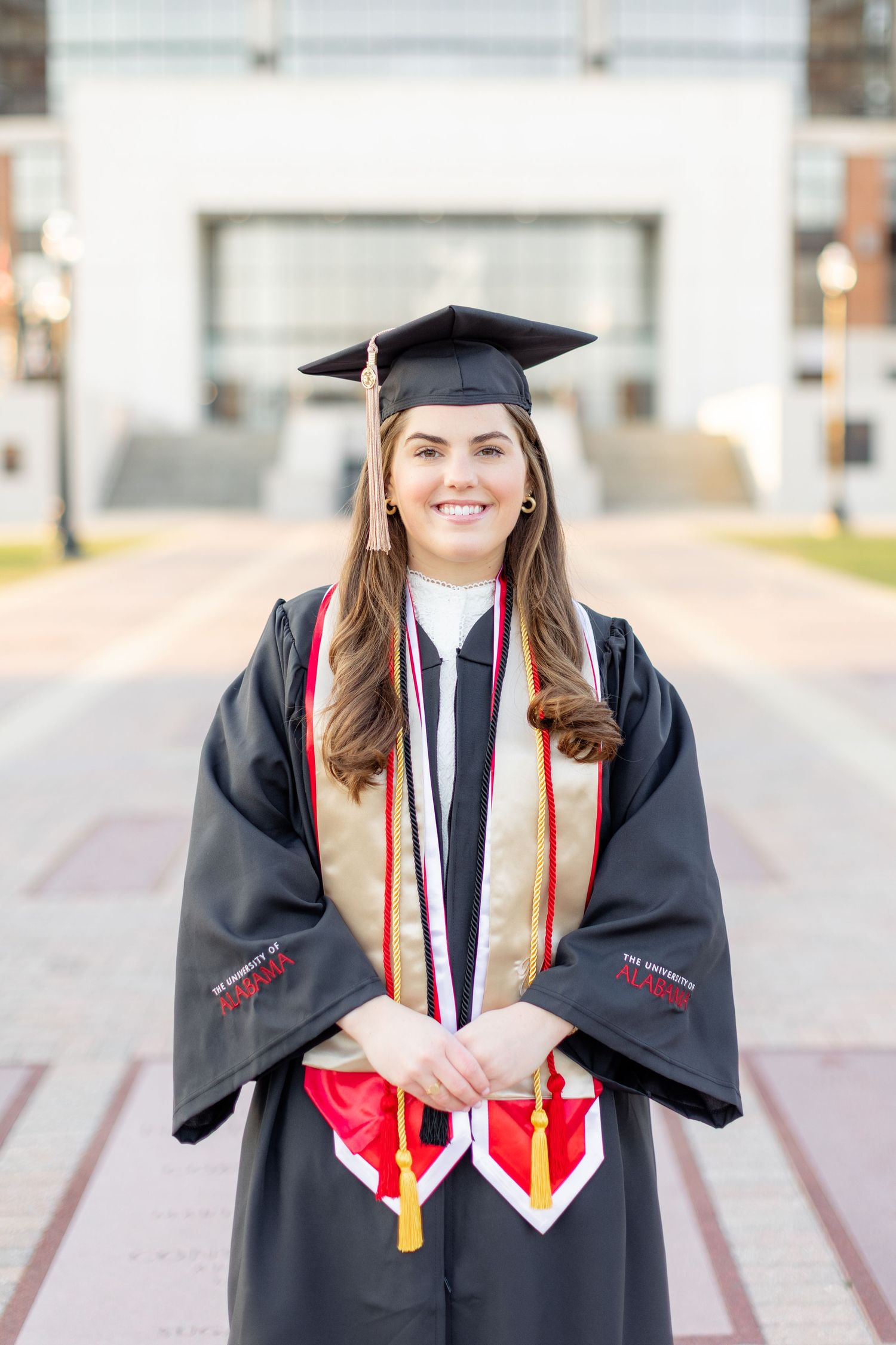 Graduate in black cap and gown with red stole smiles during outdoor graduation photo session.