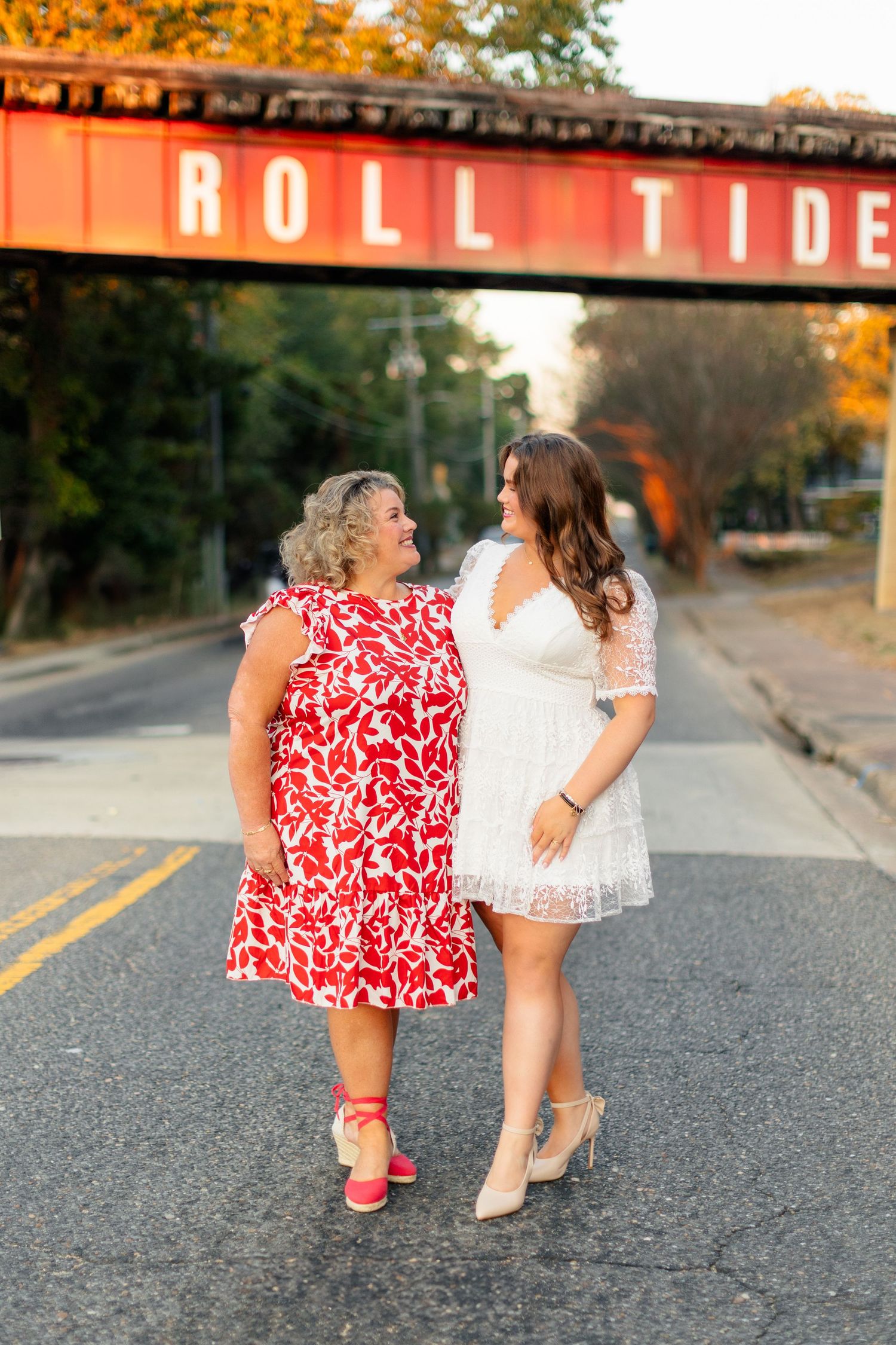 Two people in red and white dresses walking together on a road during golden hour with autumn colors in background.