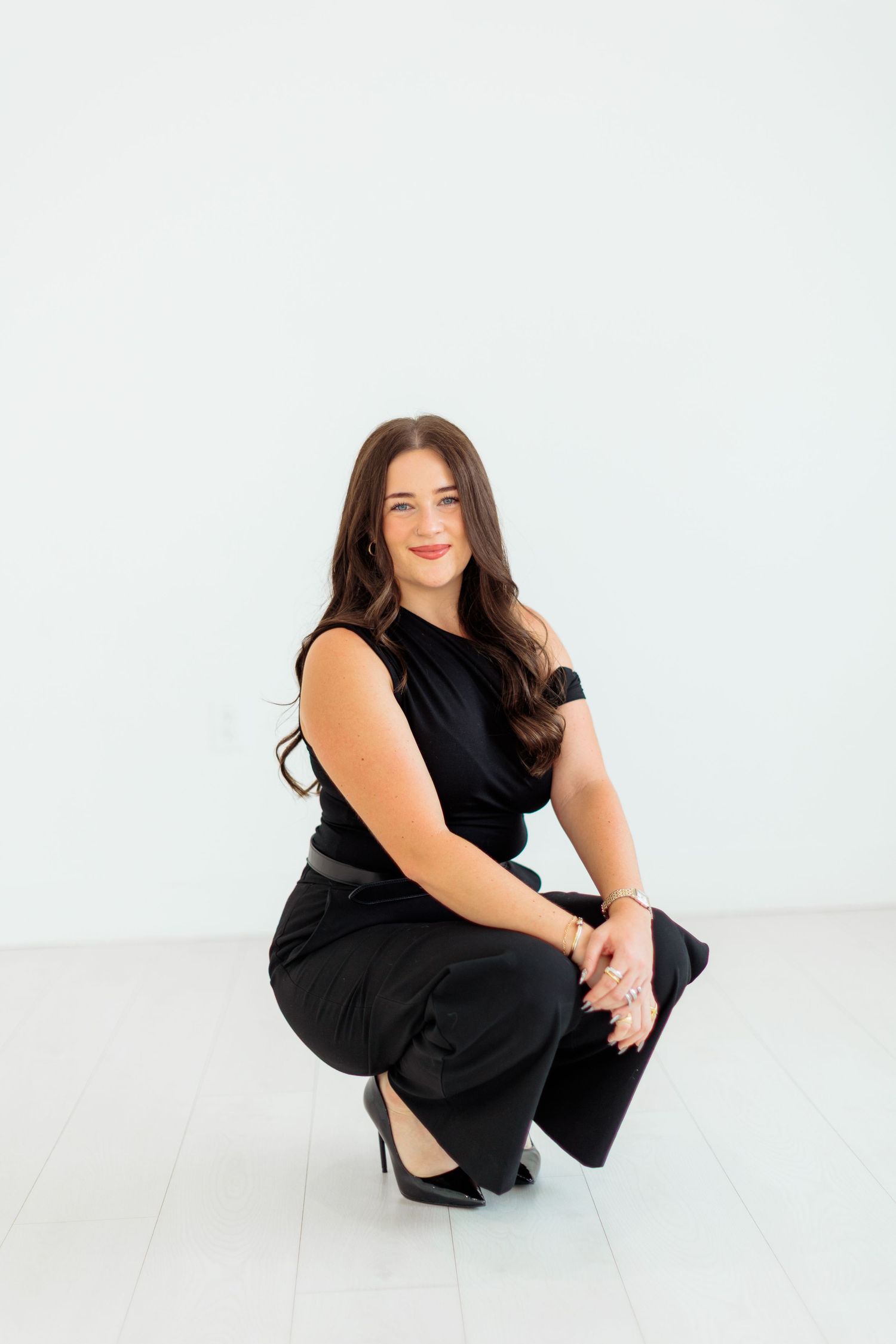 Brunette woman posing in black sleeveless outfit crouching on white floor.